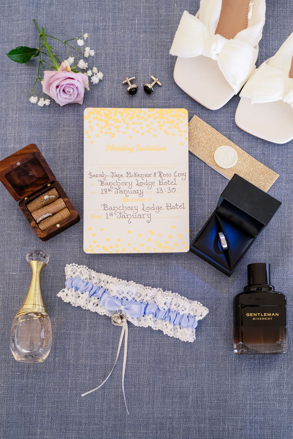 Wedding details including perfume bottles, garter, invitations, and jewelry on gray linen background.