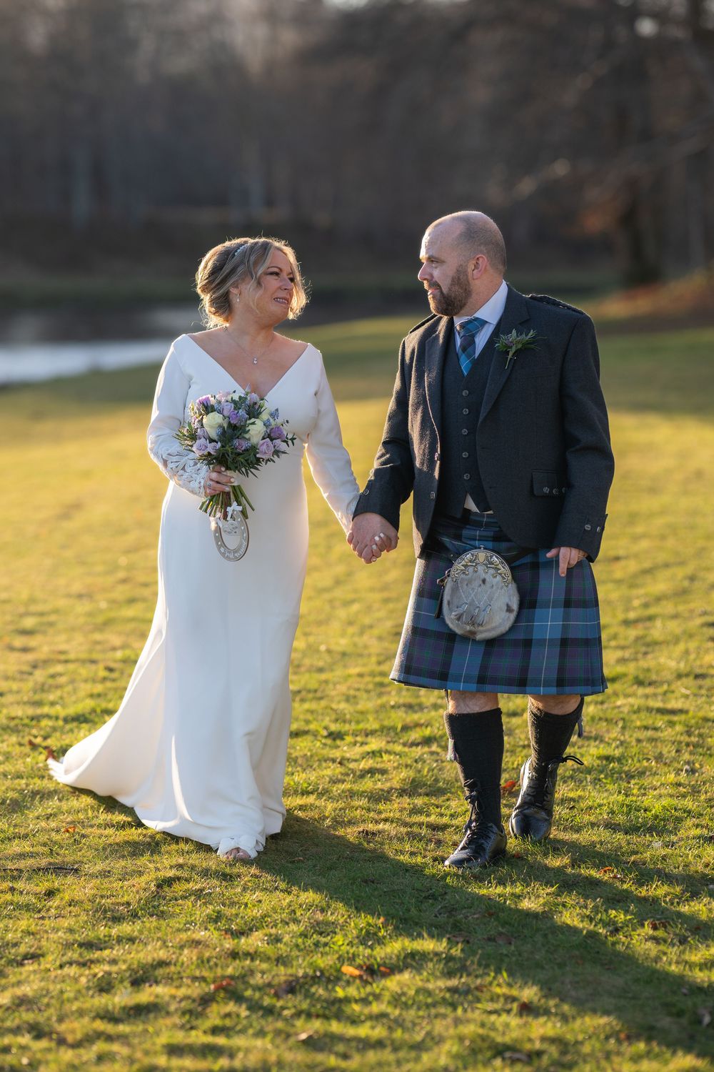 Couple in wedding attire walking on sunny grass, groom wearing traditional Scottish kilt.