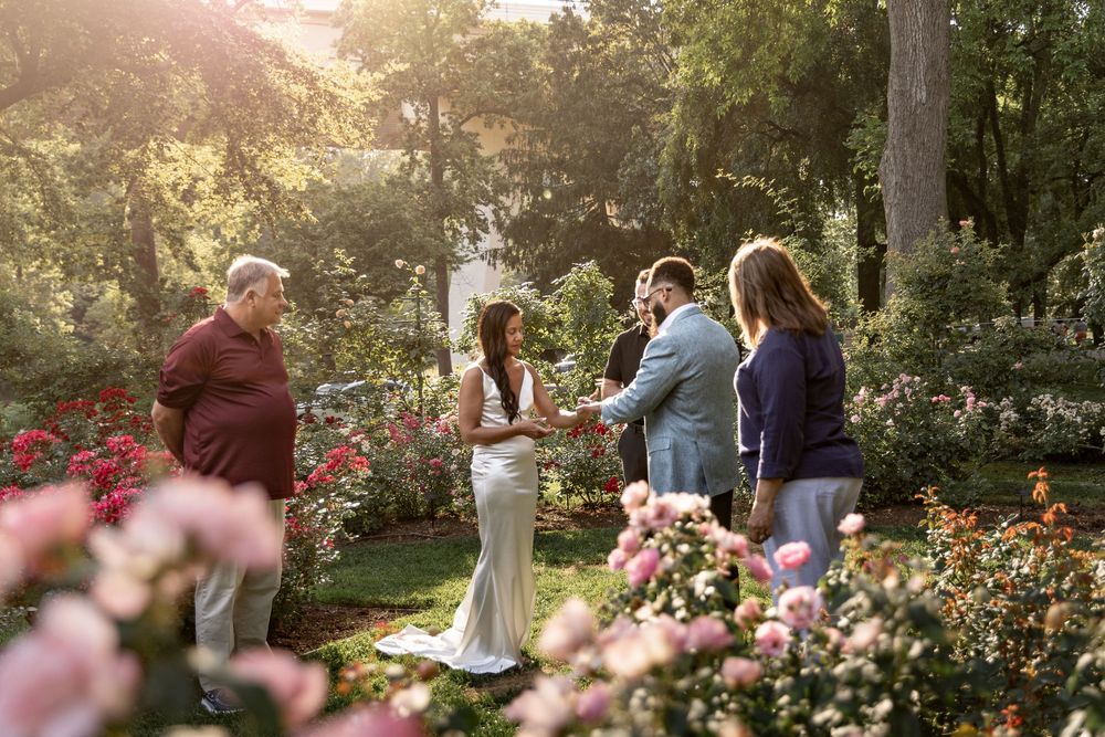 A small wedding ceremony takes place in a beautiful garden filled with blooming pink roses at sunset.