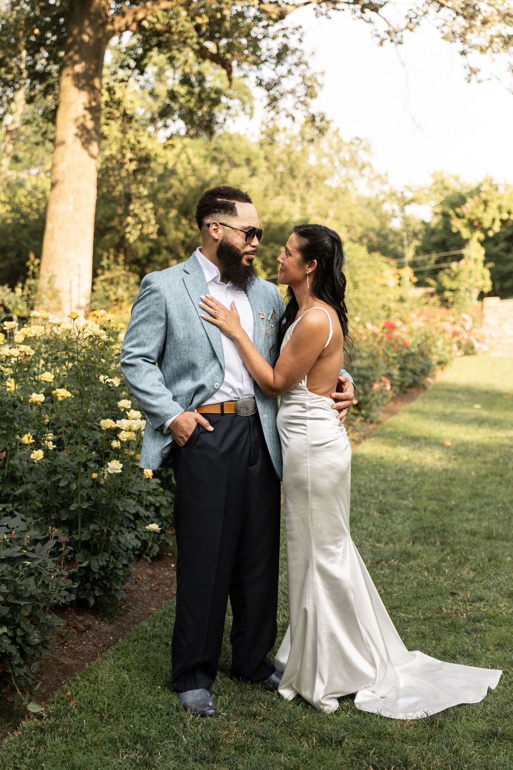 A couple shares an intimate moment in a garden setting with yellow roses and trees during a wedding photoshoot.