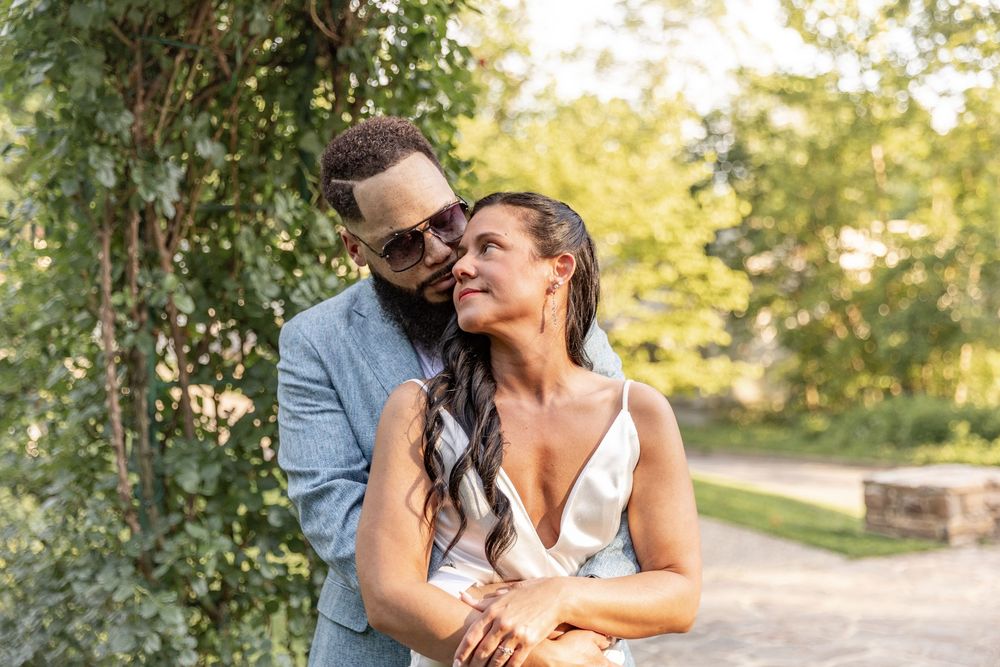 A romantic couple embraces outdoors in a garden setting with natural sunlight filtering through trees.