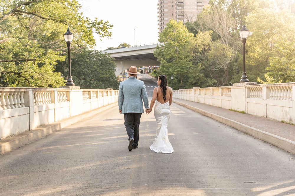 A couple in wedding attire walks along a scenic bridge path during golden hour with sunlit trees in the background.
