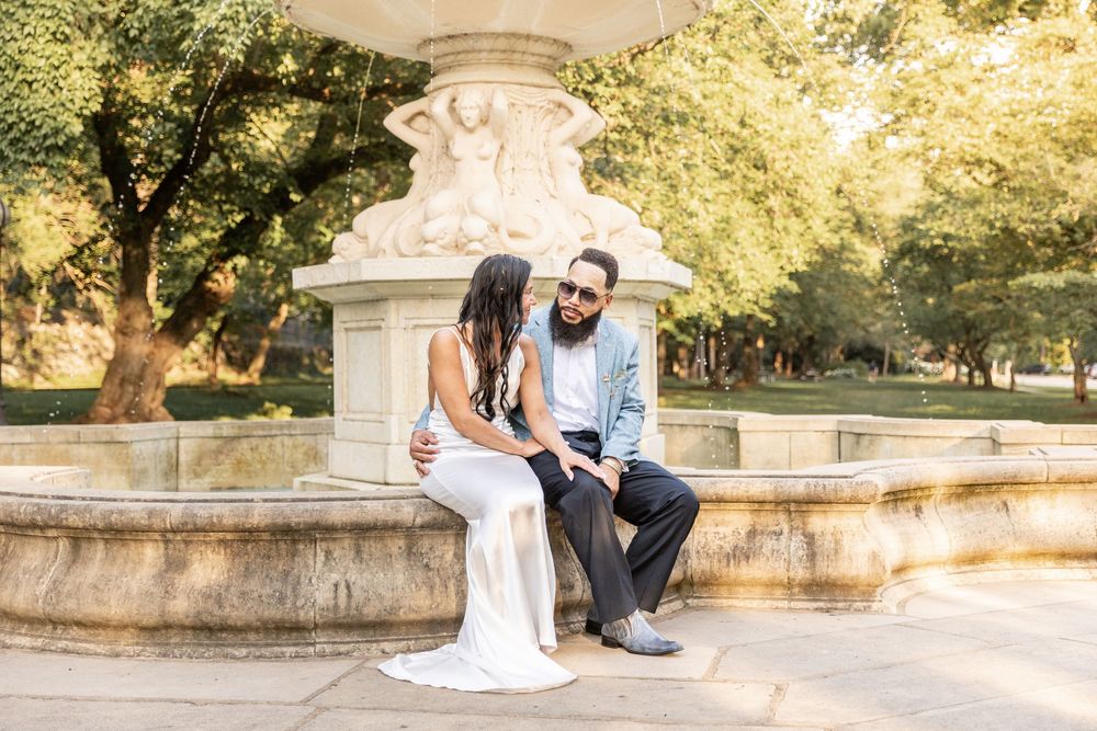 A couple sits together on stone steps in a sunlit park setting during a romantic outdoor photoshoot.