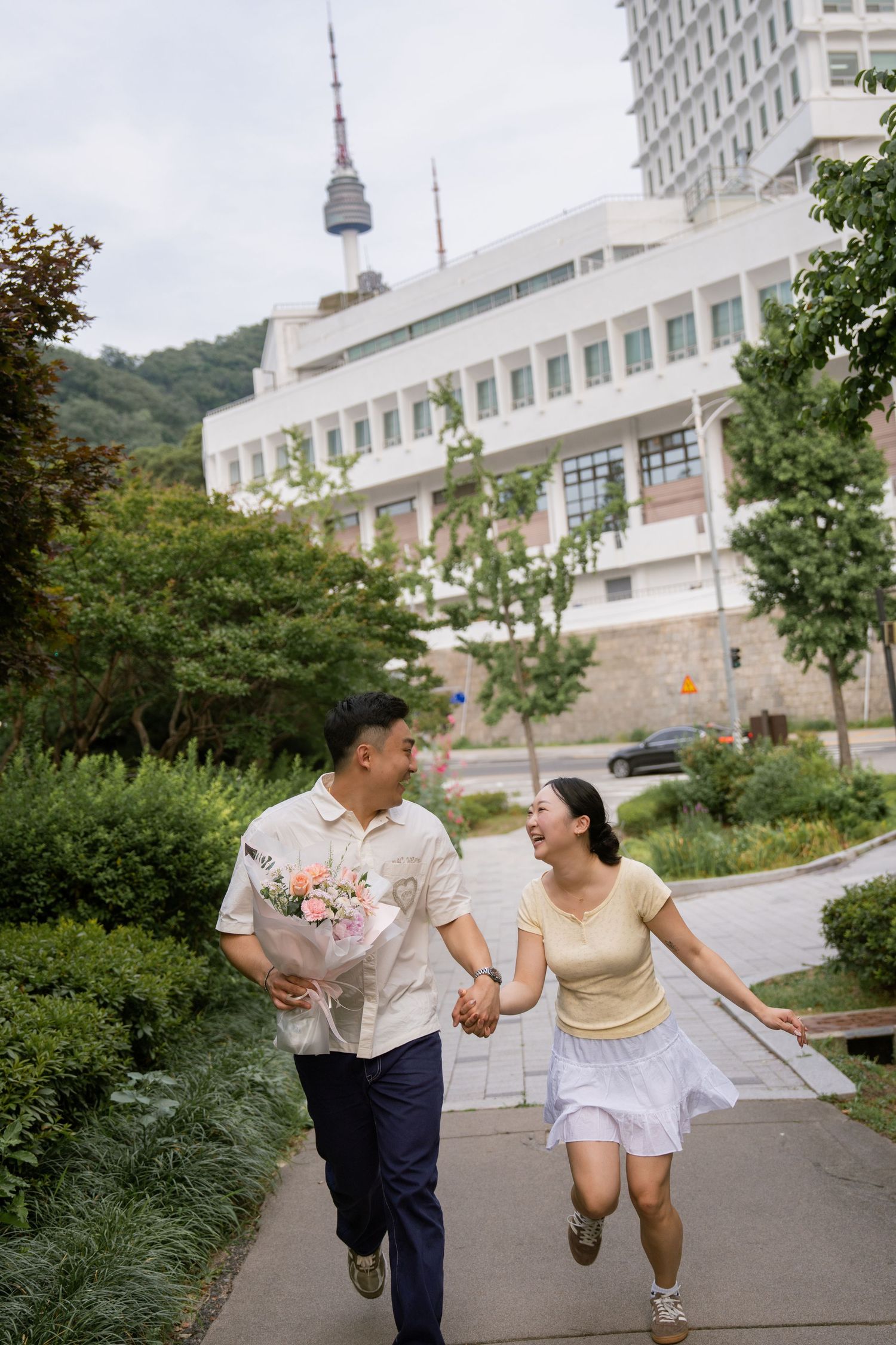 A couple walks hand in hand along a winding path outside a modern building complex on a sunny day.