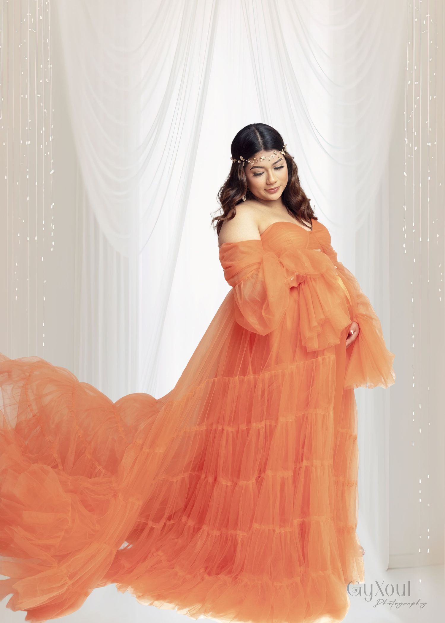 A flowy orange tulle gown with multiple tiers creates a dreamy silhouette against a white curtained backdrop.