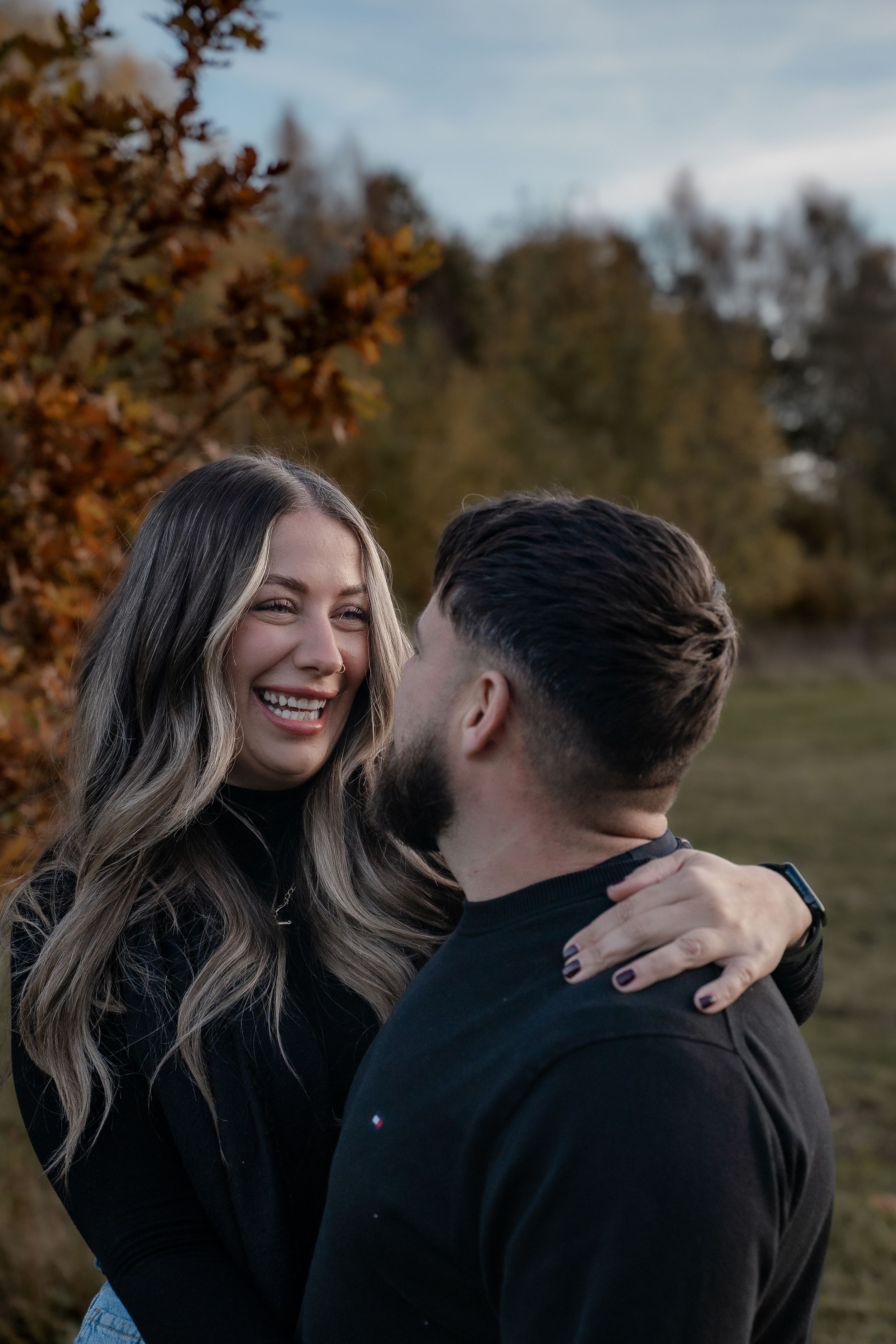 A couple wearing black outfits share intimate moments and laughter during an autumn outdoor photoshoot.