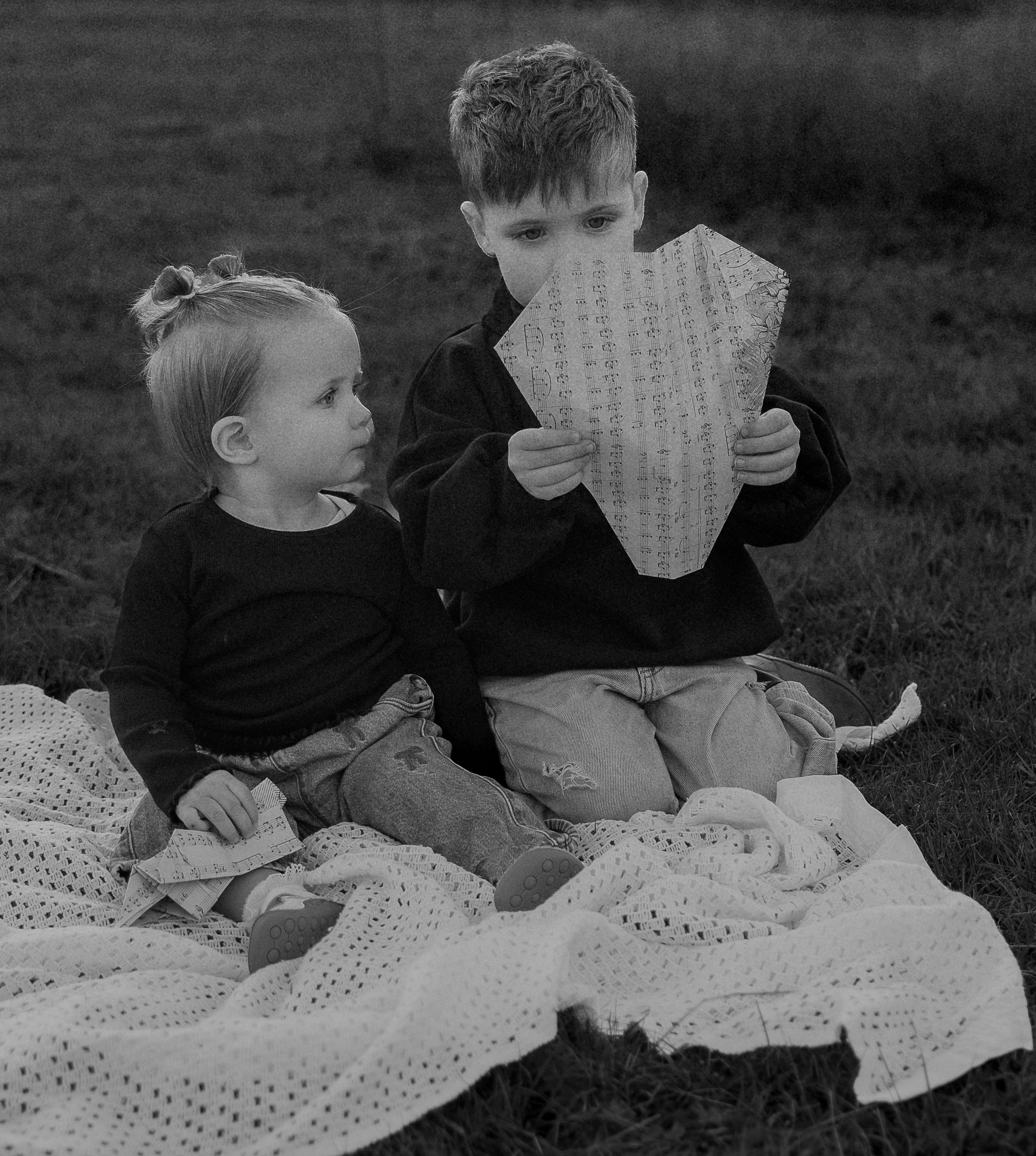 Two children sit on a white blanket outdoors while looking at handwritten notes in a black and white photograph.