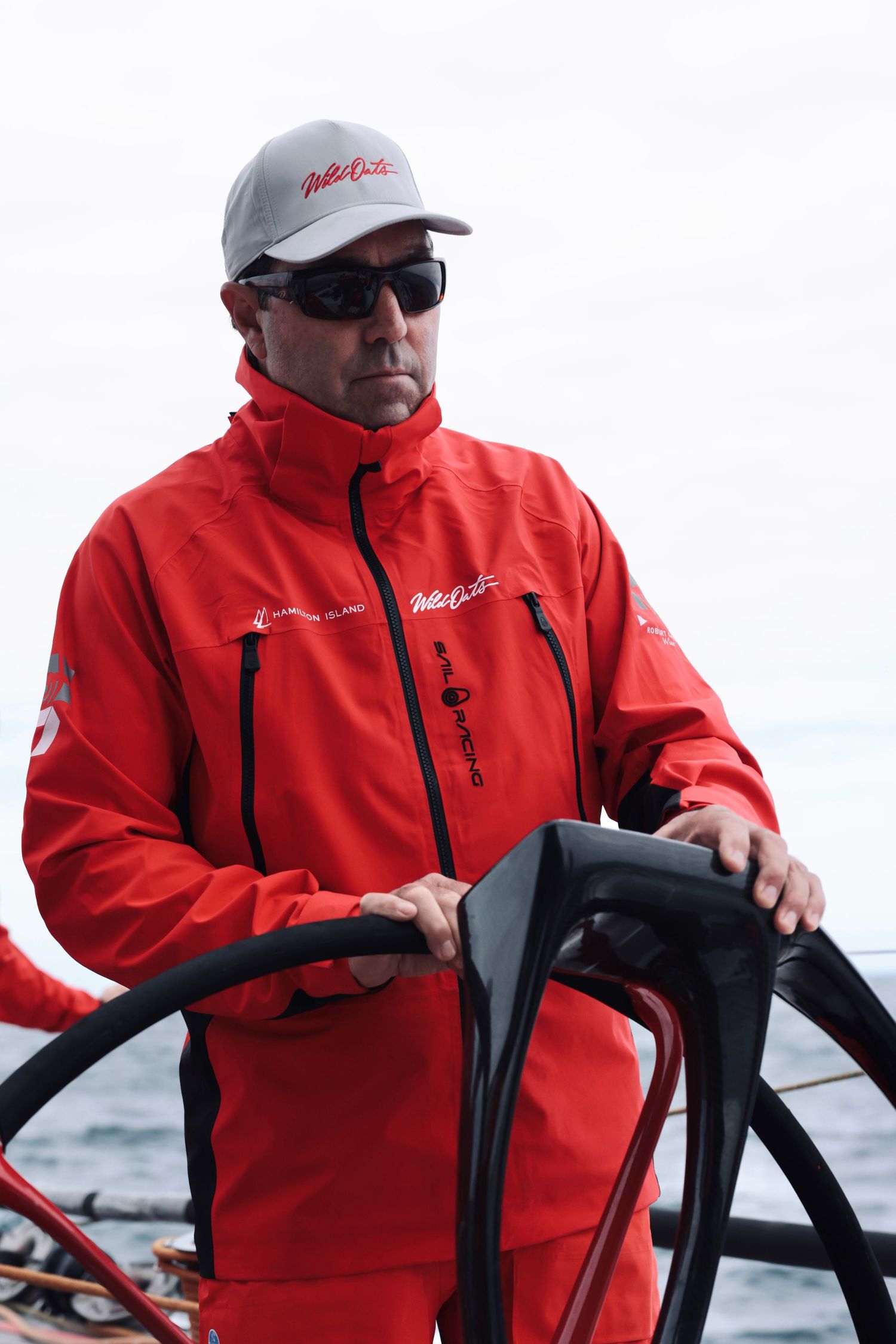 Crew member in red sailing jacket at helm of racing yacht in overcast conditions.