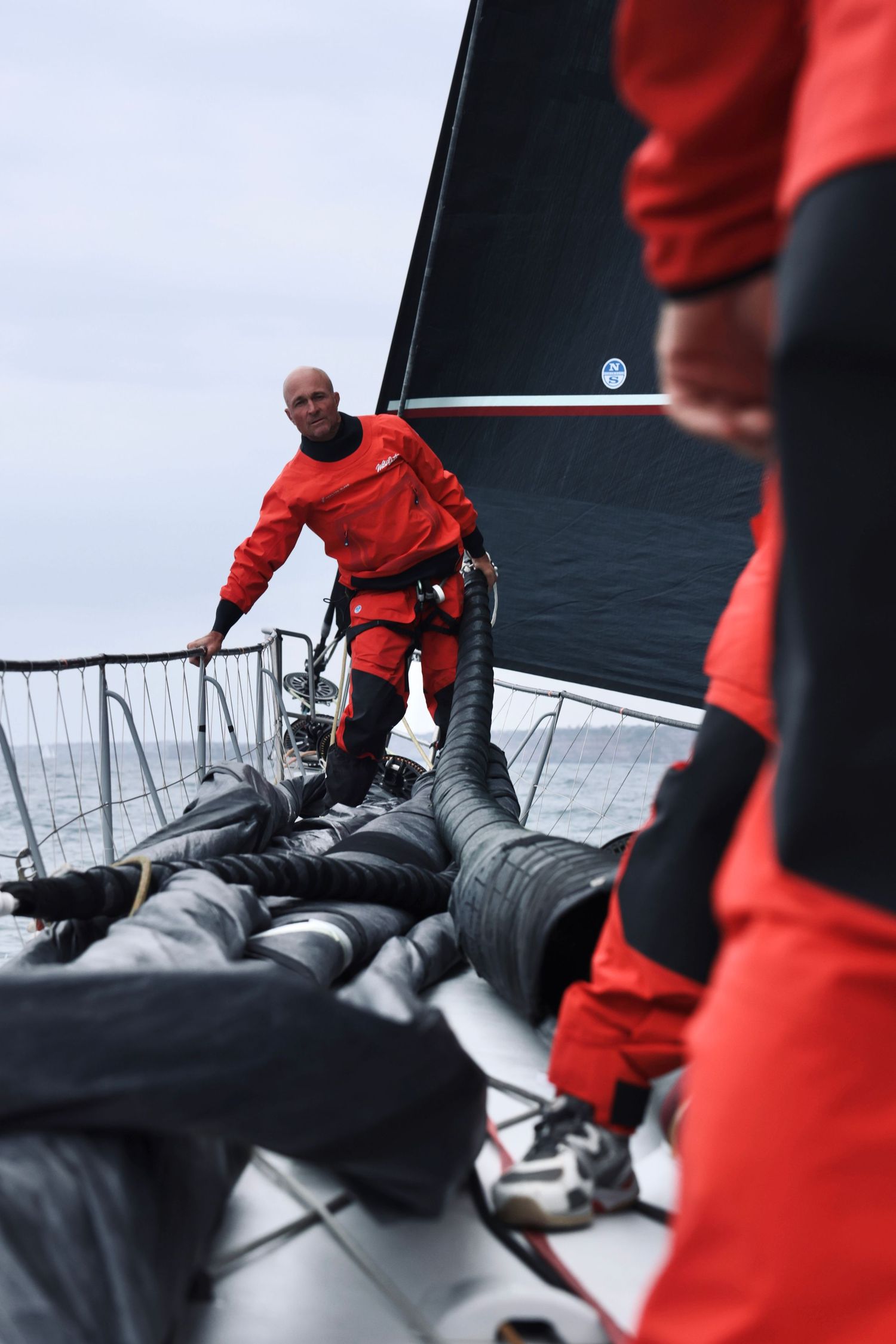 Crew member in red safety gear navigating across deck of racing yacht at sea.
