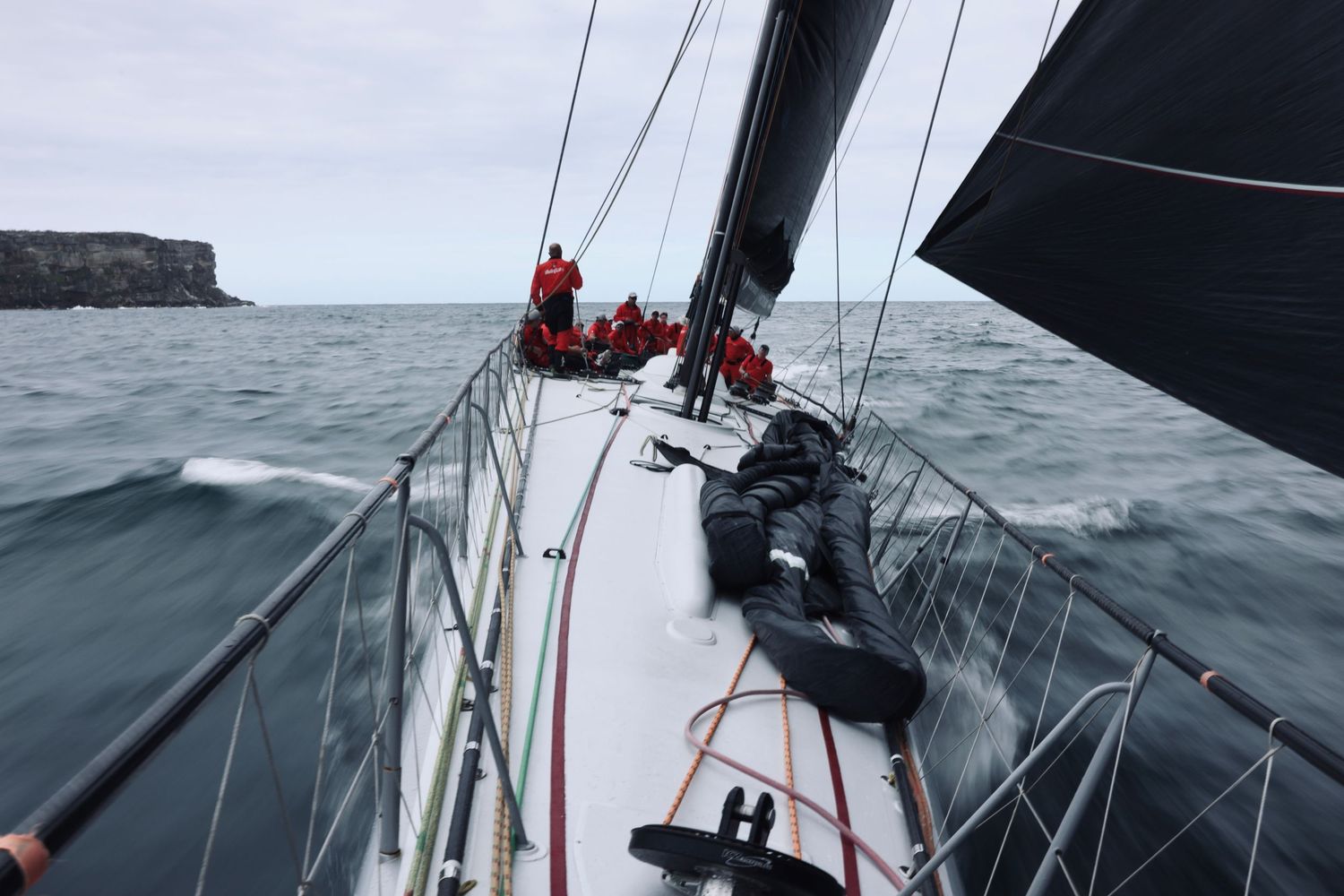 View along deck of racing yacht with crew in red gear sailing through choppy waters.