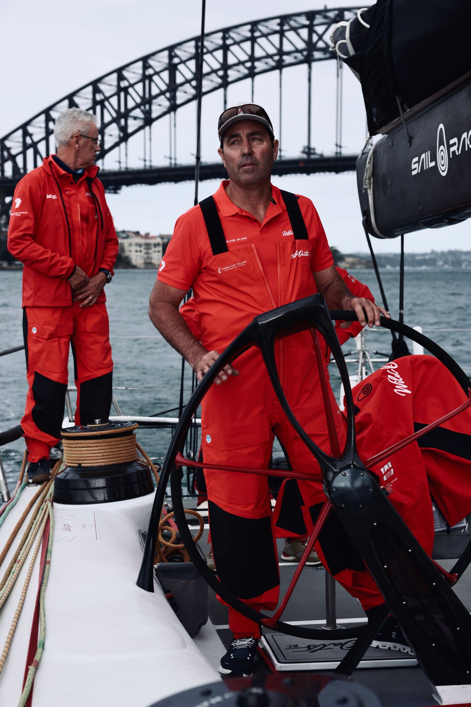 Crew members in red sailing gear operate equipment on deck with Sydney Harbour Bridge in background.