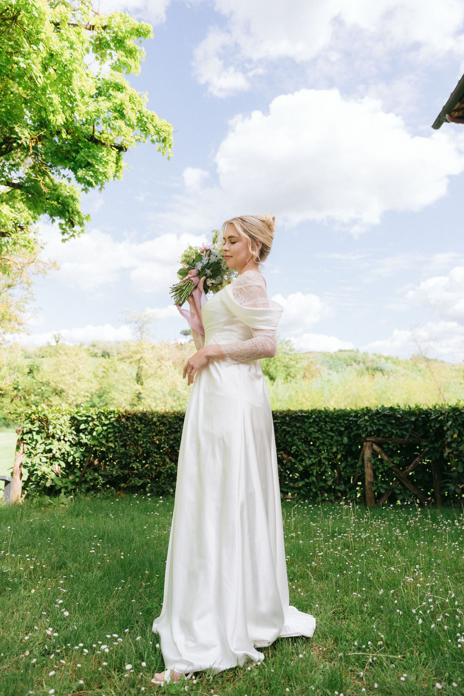 Bride in flowing white dress twirls in a sunlit meadow against blue sky.