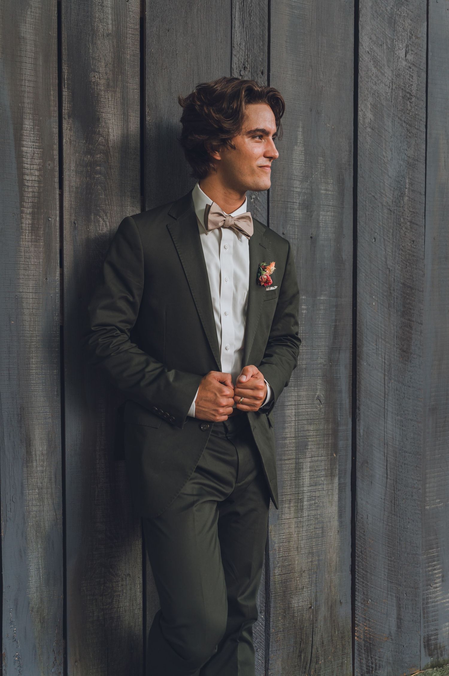 Person in dark suit and bowtie poses against weathered wooden wall background.