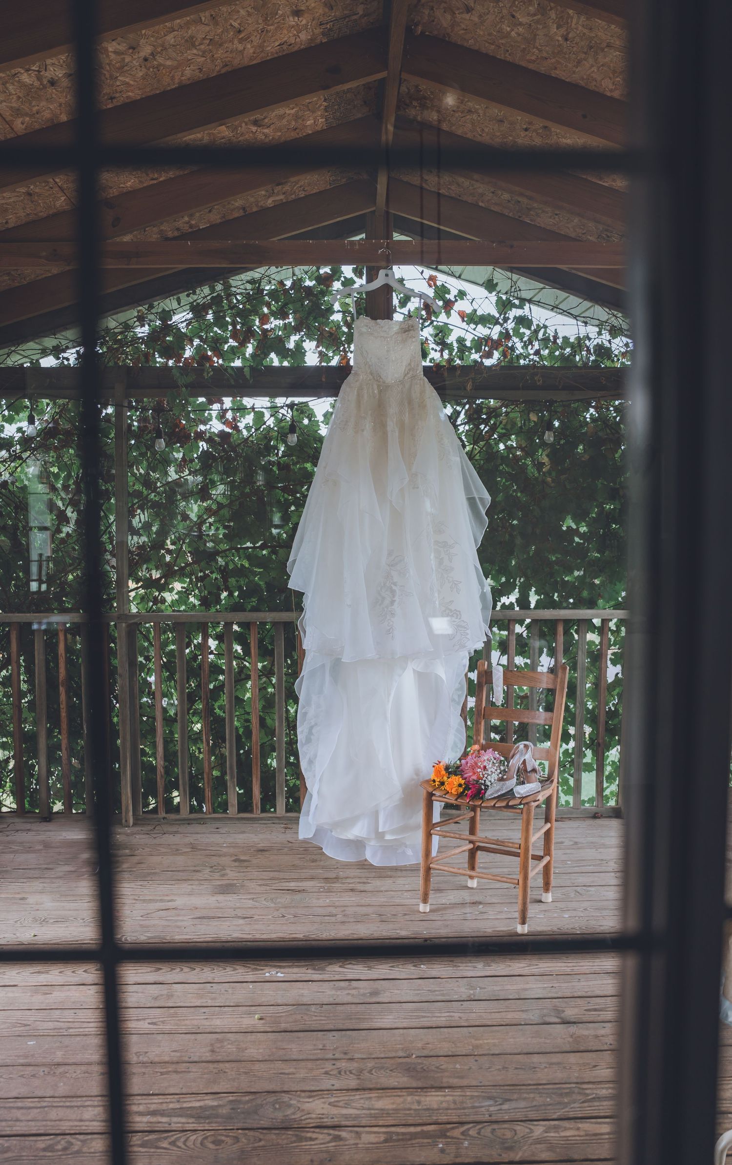 White wedding dress hangs elegantly on wooden deck railing with forest backdrop and rustic wooden chairs.