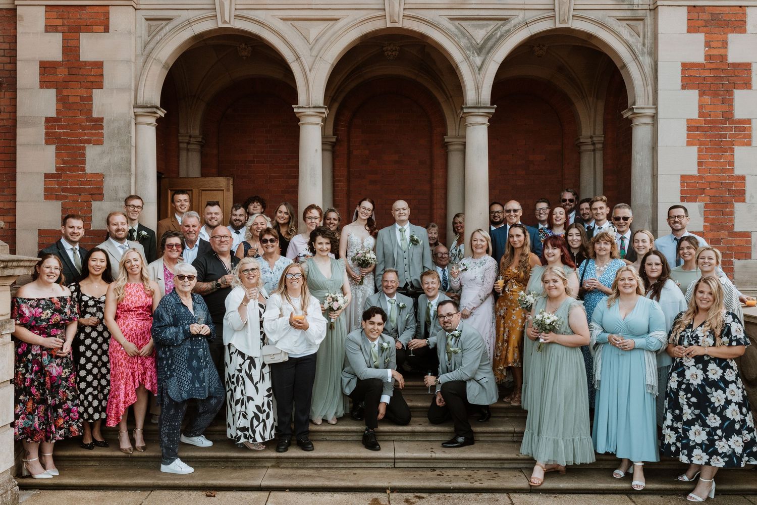 Large wedding party poses together in front of a brick building with arched columns and windows.