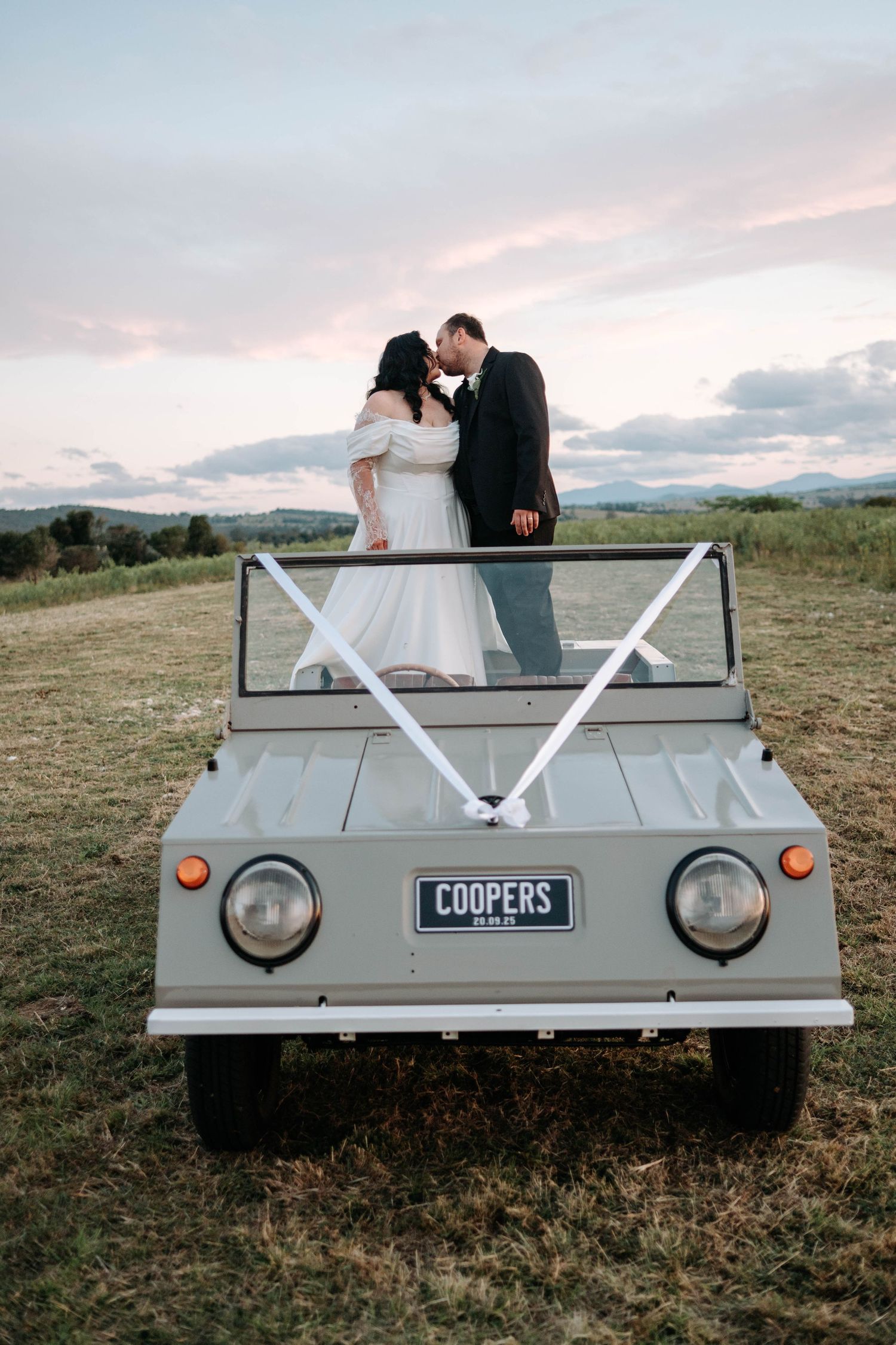 Newlywed couple sits in back of vintage Coopers car during sunset photo session in countryside field.