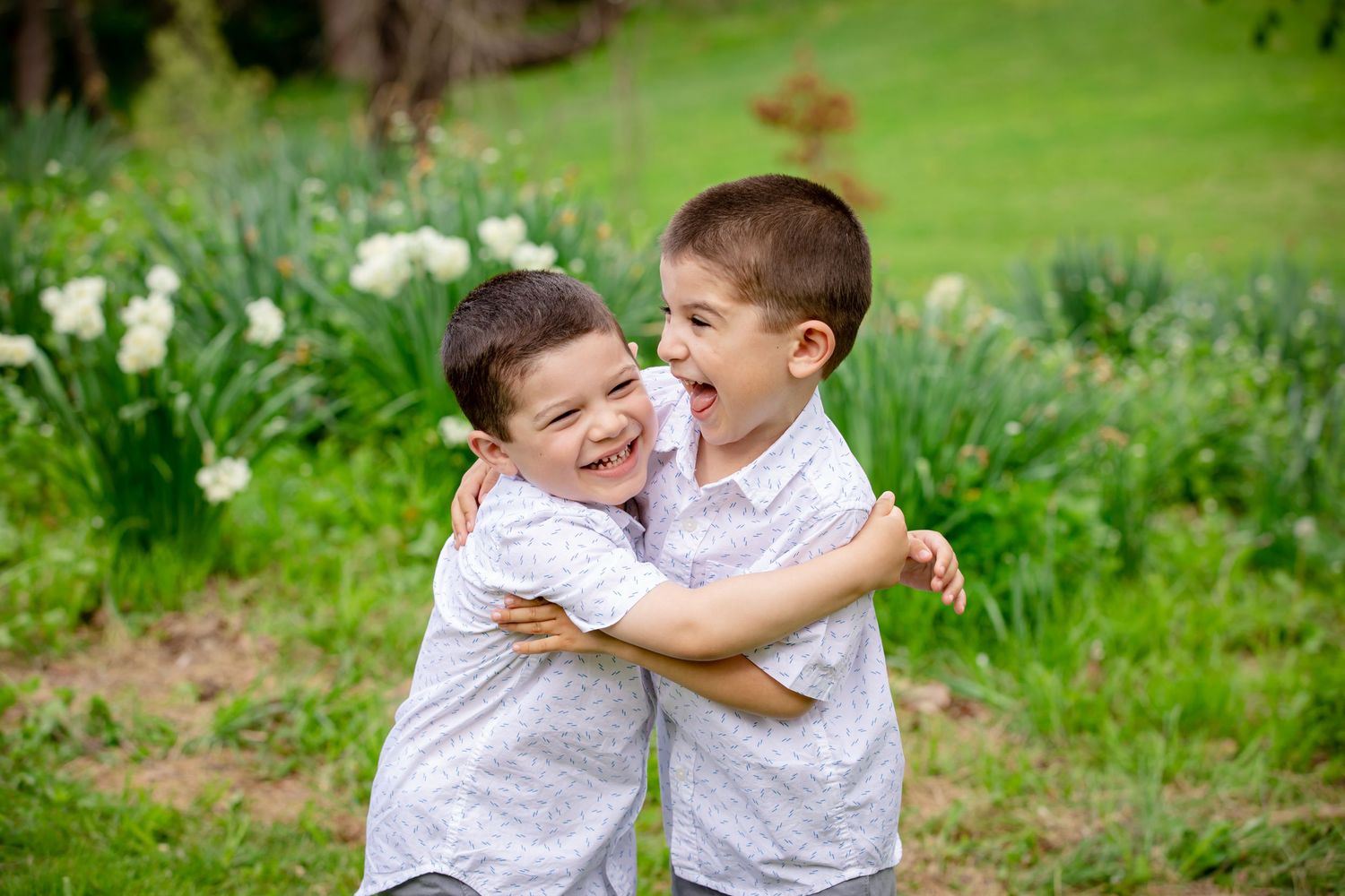 Two children in white shirts play and laugh together on green grass in a sunny outdoor setting.