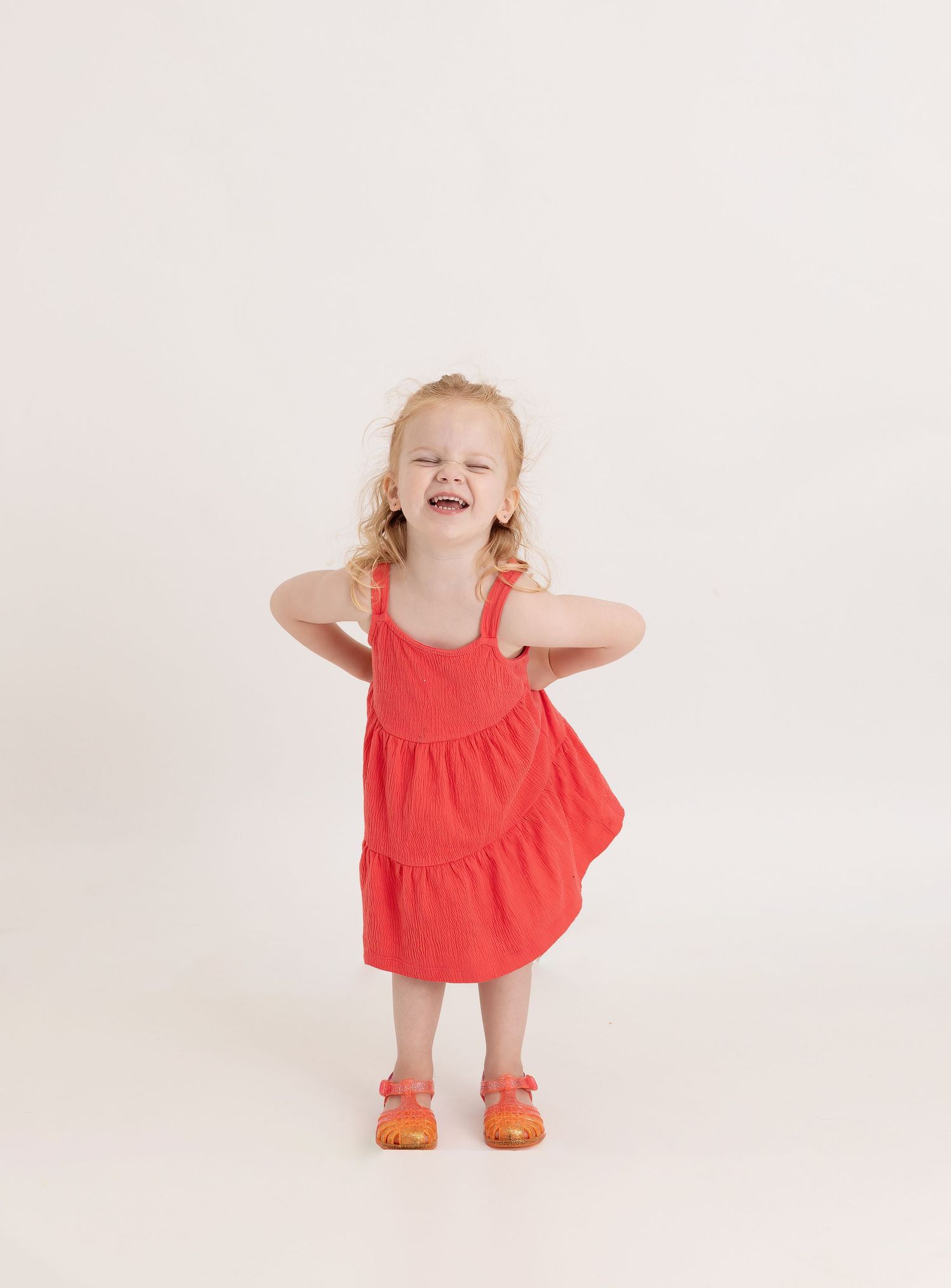 A small child wearing a coral dress and orange shoes twirls and dances in a white studio setting.