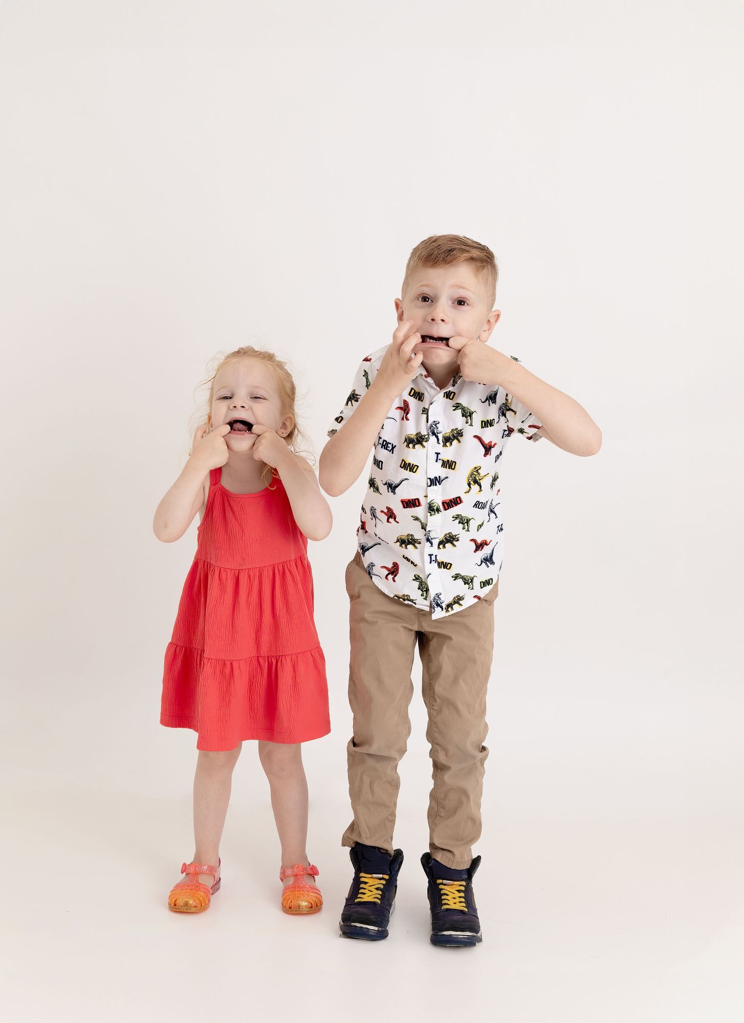 Two young children in casual outfits stand together on a white background showing playful expressions.