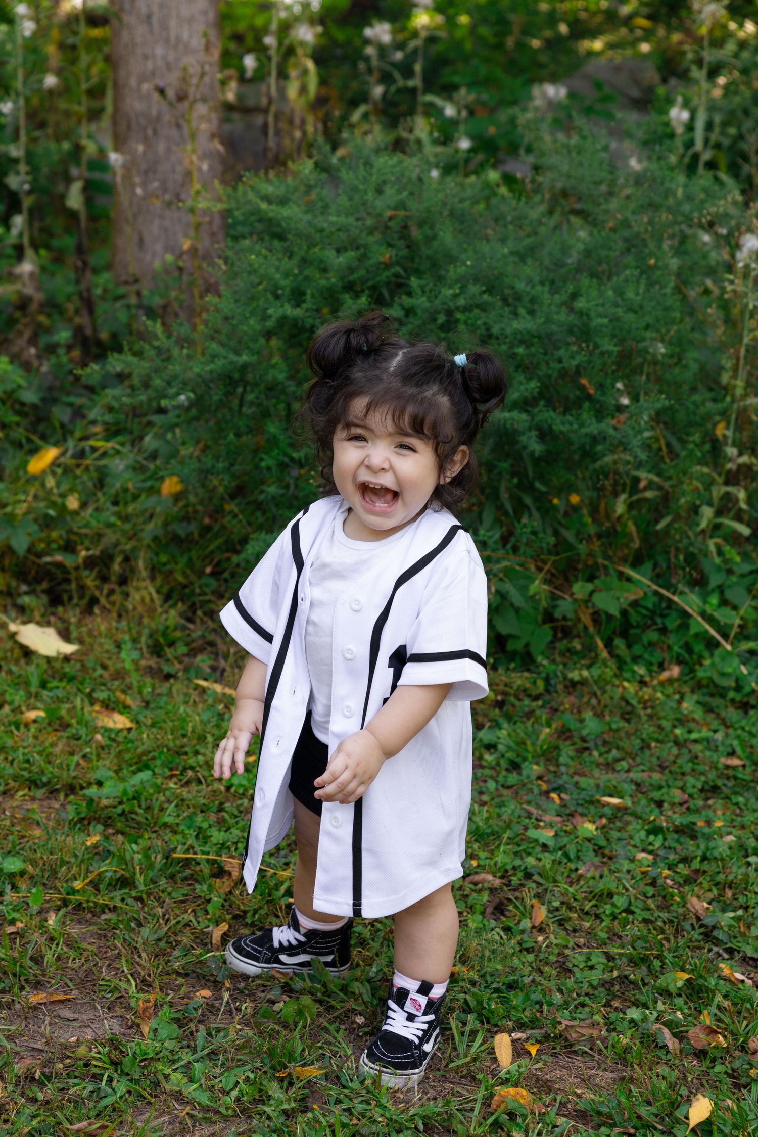 A child in a white shirt and black pants plays outdoors among trees and fallen leaves in a natural setting.