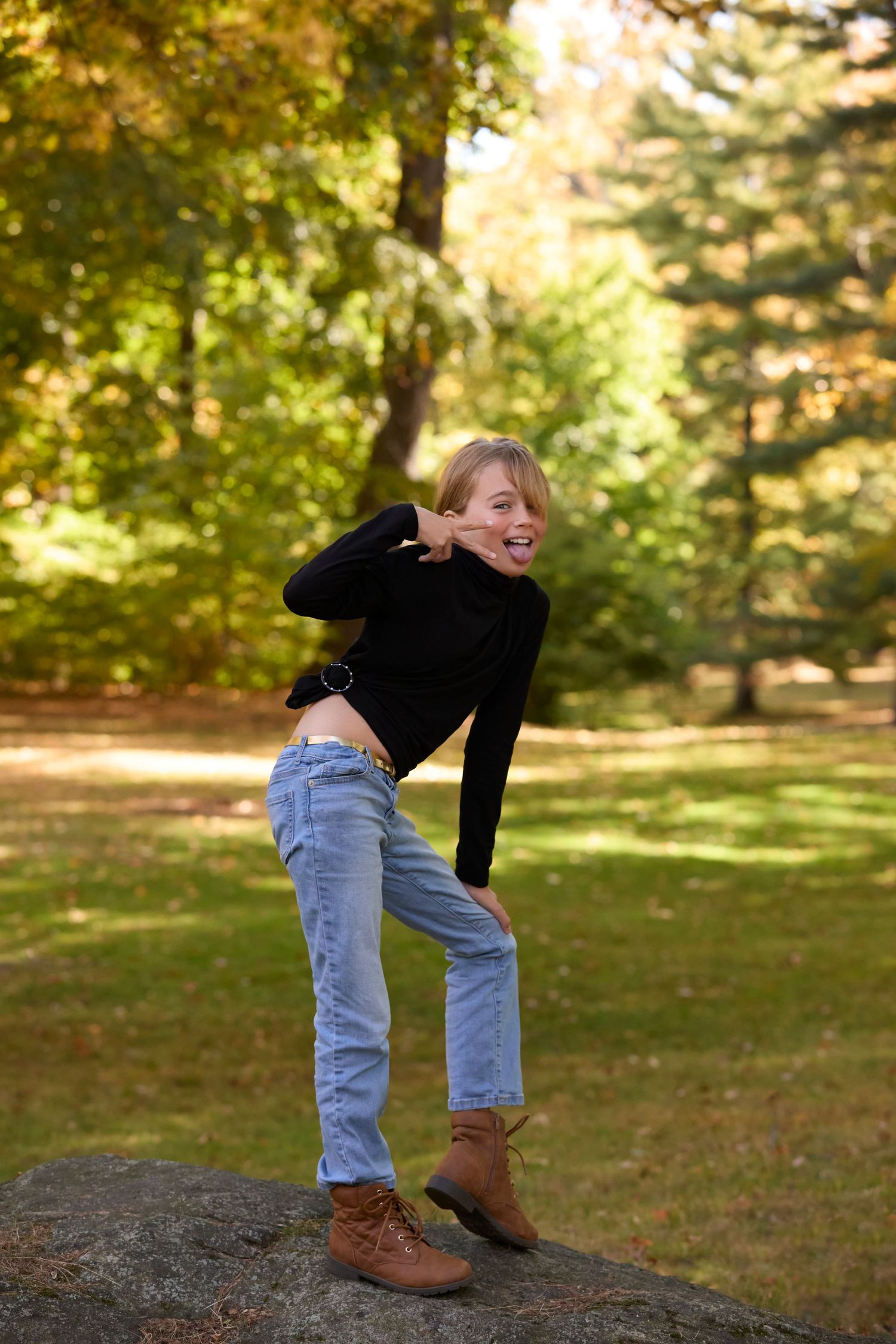 Person in jeans and brown boots bending over in a park setting with autumn trees in the background.