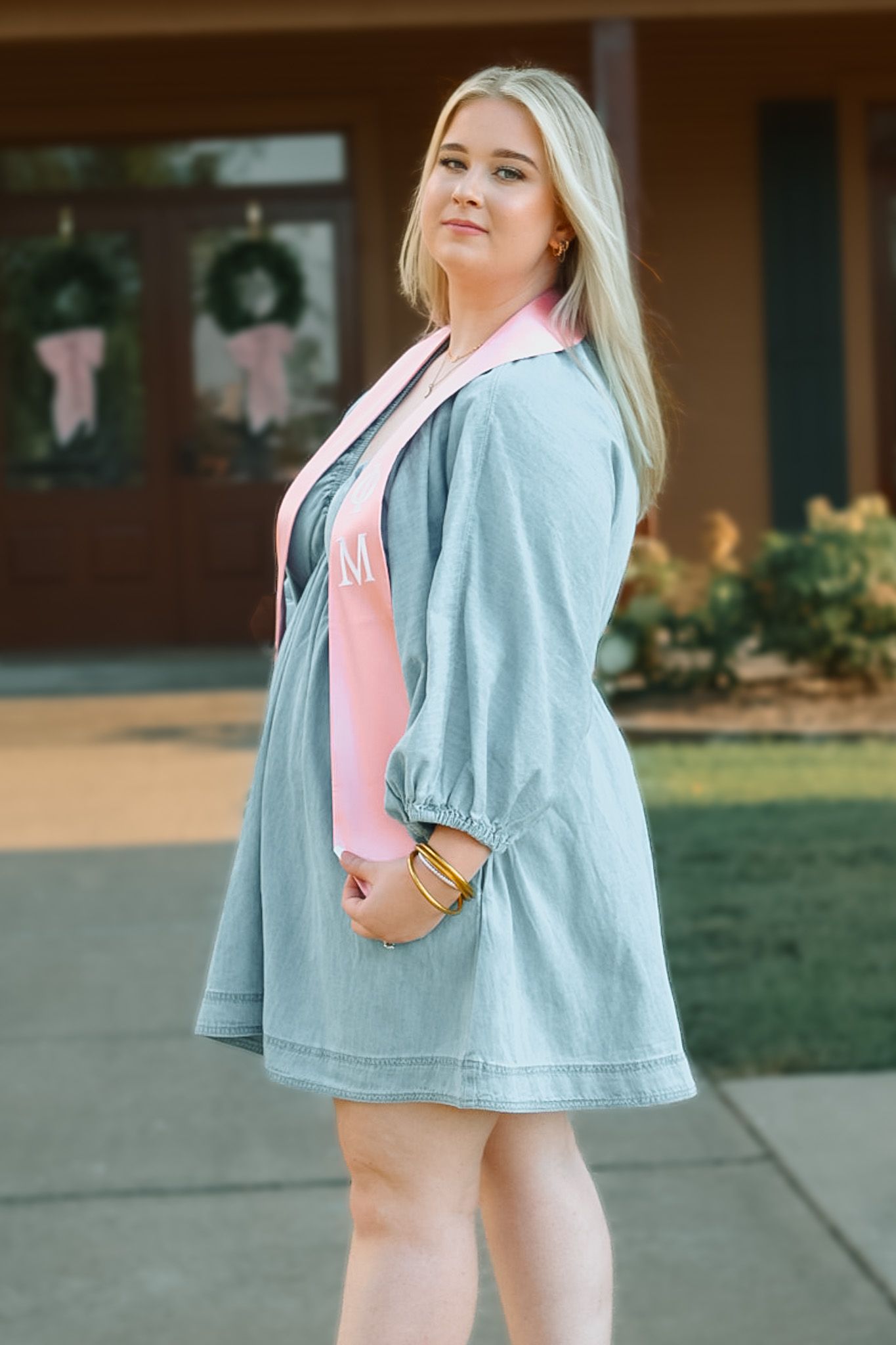 A woman in a light blue denim jacket and pink dress poses confidently outdoors.