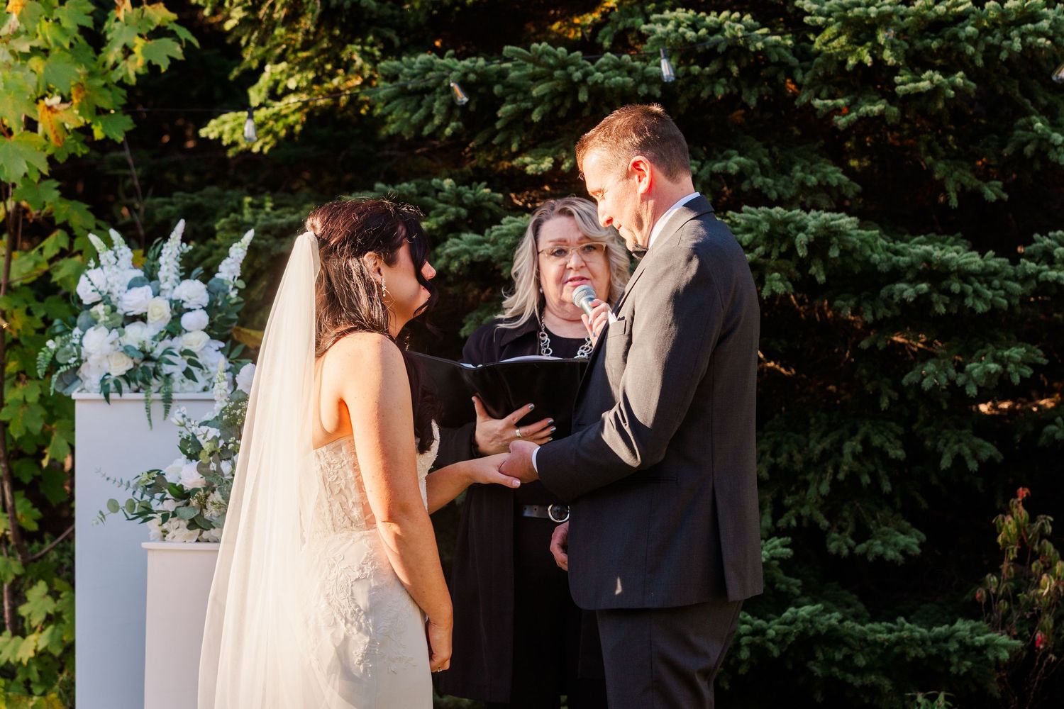 A wedding ceremony takes place outdoors with an officiant and couple standing before white floral arrangements and greenery.