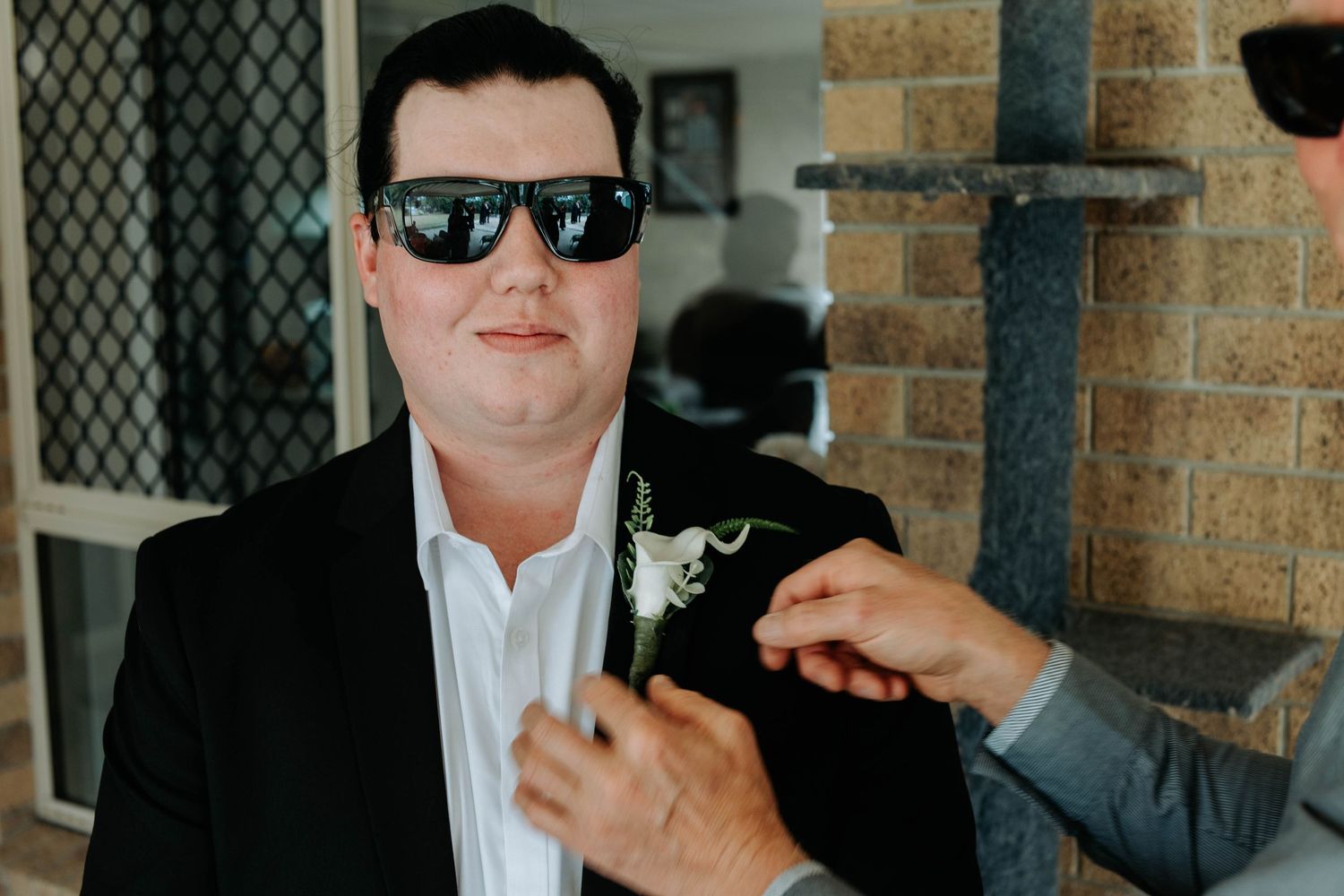 Three people in dark suits and sunglasses stand against a brick wall in a formal setting.