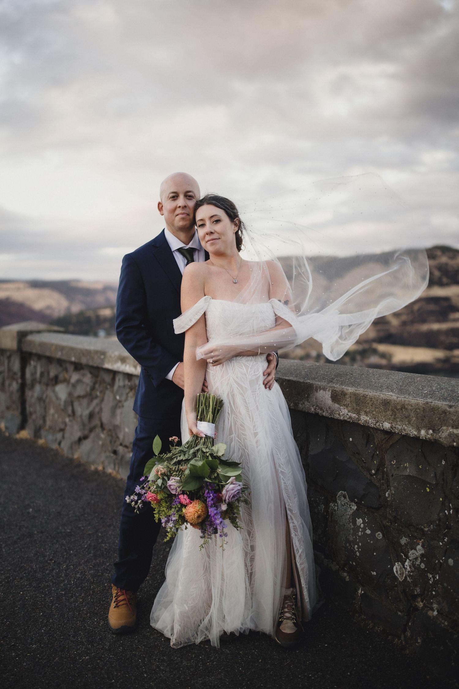 A romantic wedding couple embraces on a stone wall against a dramatic cloudy mountain landscape.