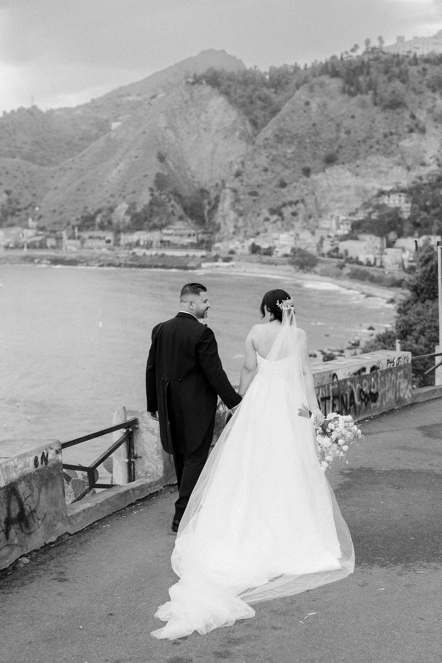 A bride and groom walk along a scenic coastal path with mountains and water views in black and white photography.