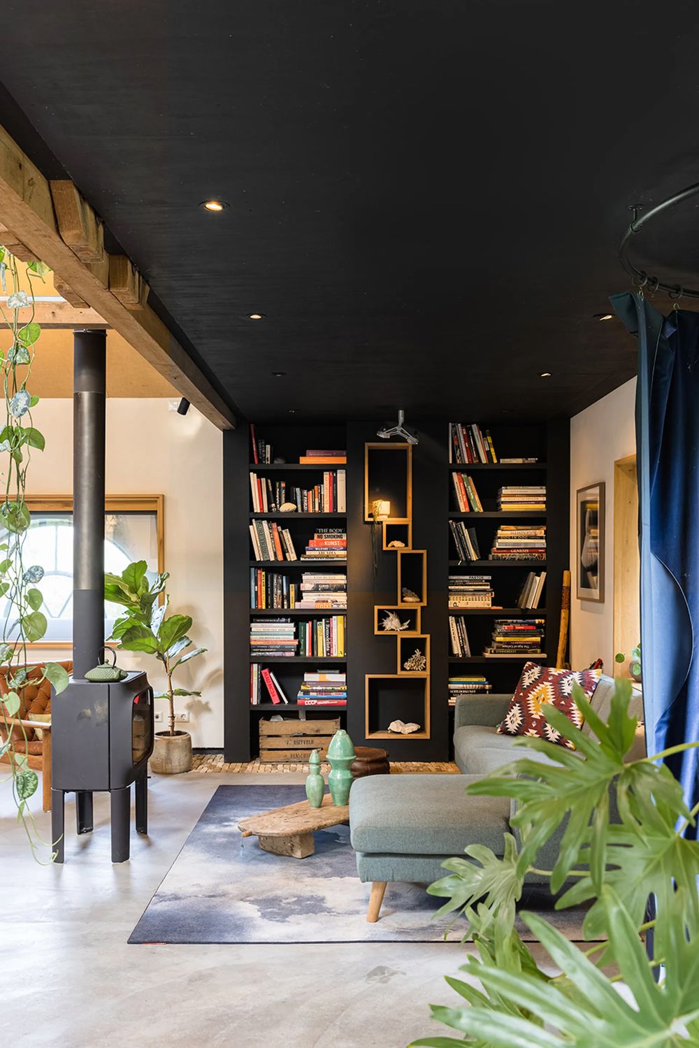 Modern home library space with black ceiling, geometric wooden shelving, indoor plants, and natural lighting.