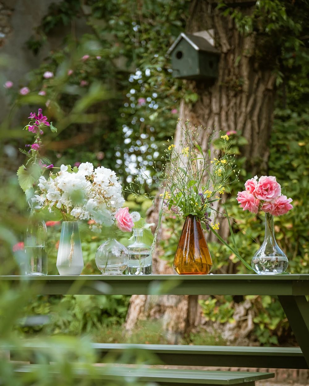 Delicate pink and white flowers arranged in glass vases and bottles on an outdoor table in a garden setting.