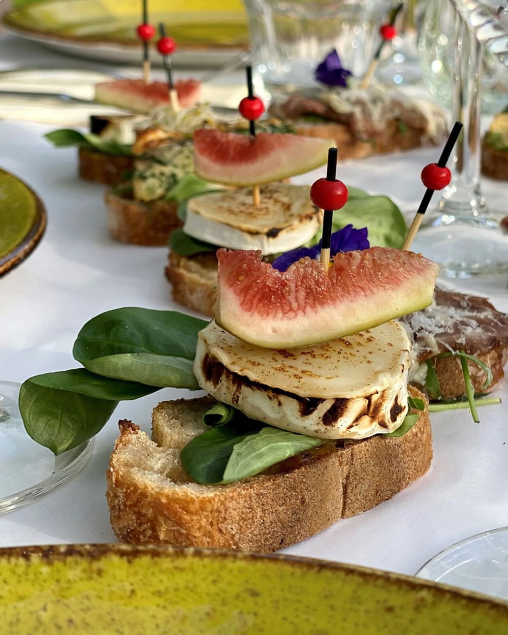 Gourmet open-faced sandwiches with figs, spinach leaves and garnishes on artisanal bread slices arranged on white plates.
