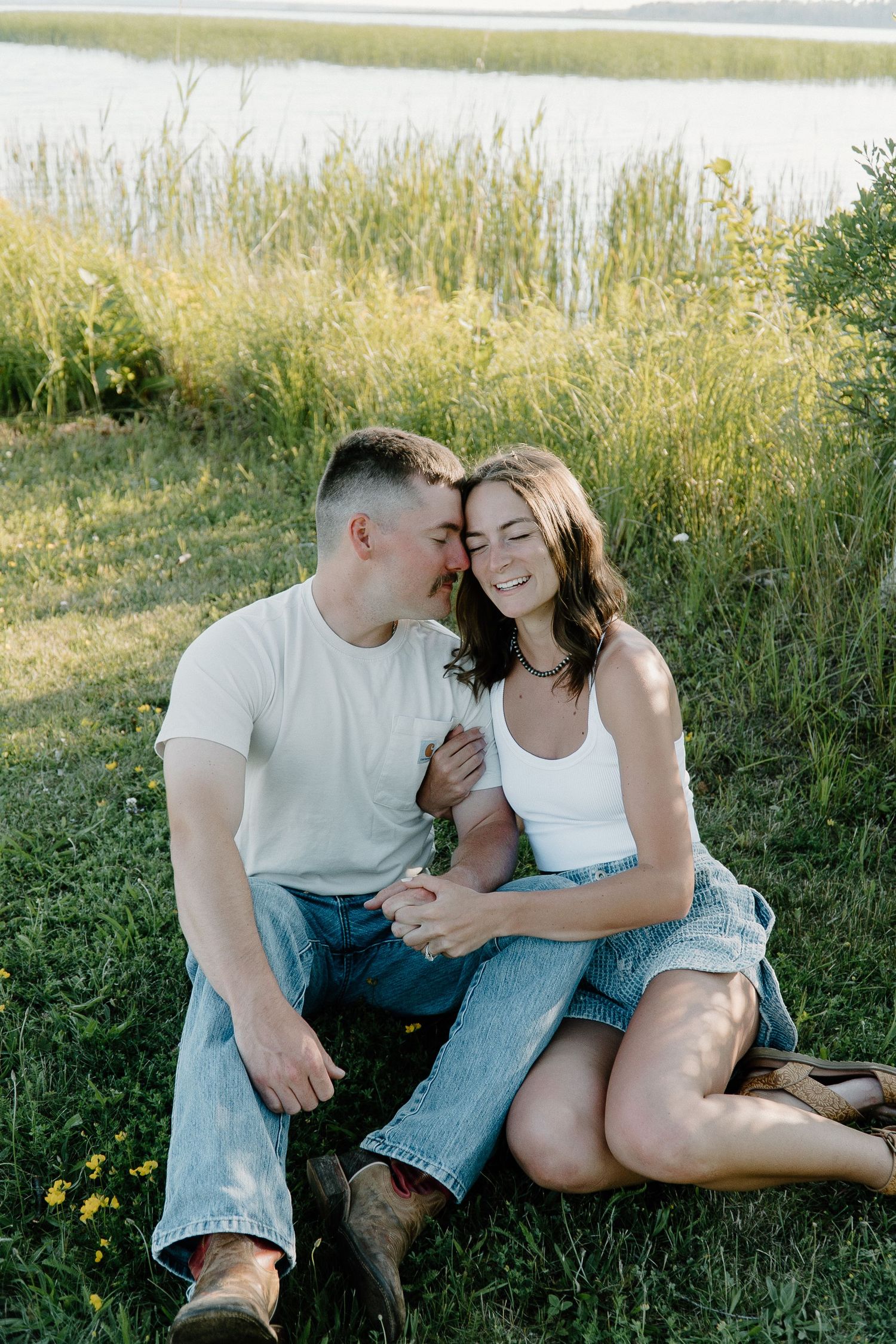 A couple sits together in a grassy meadow during a romantic summer photoshoot.