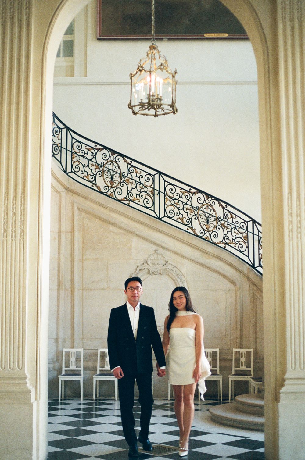 Bride and groom stand beneath ornate archways and grand staircase in classic mansion interior. Musee Rodin photoshoot.