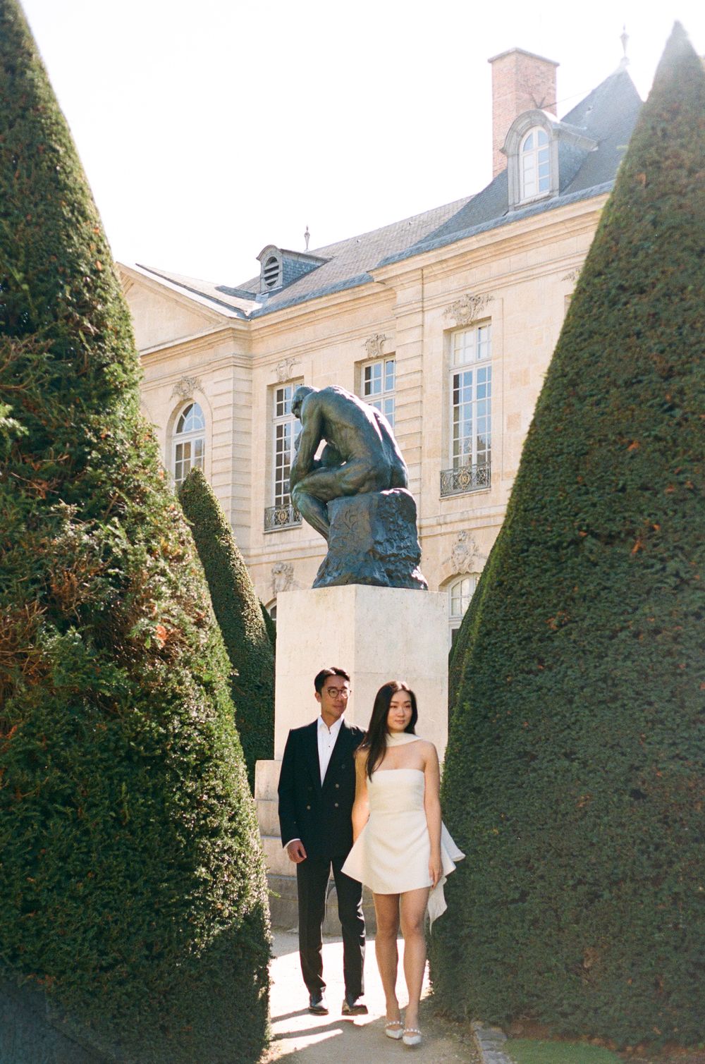 Couple walks through garden path lined with perfectly manicured tall topiary trees. Paris pre wedding photoshoot on film photography.