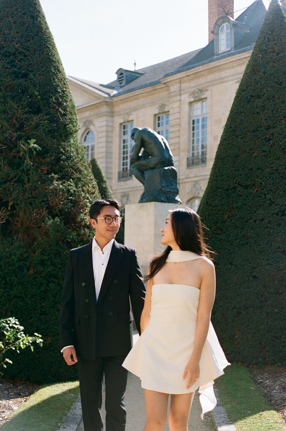 A couple in wedding attire walks through a garden with manicured hedges and a historic building in the background.