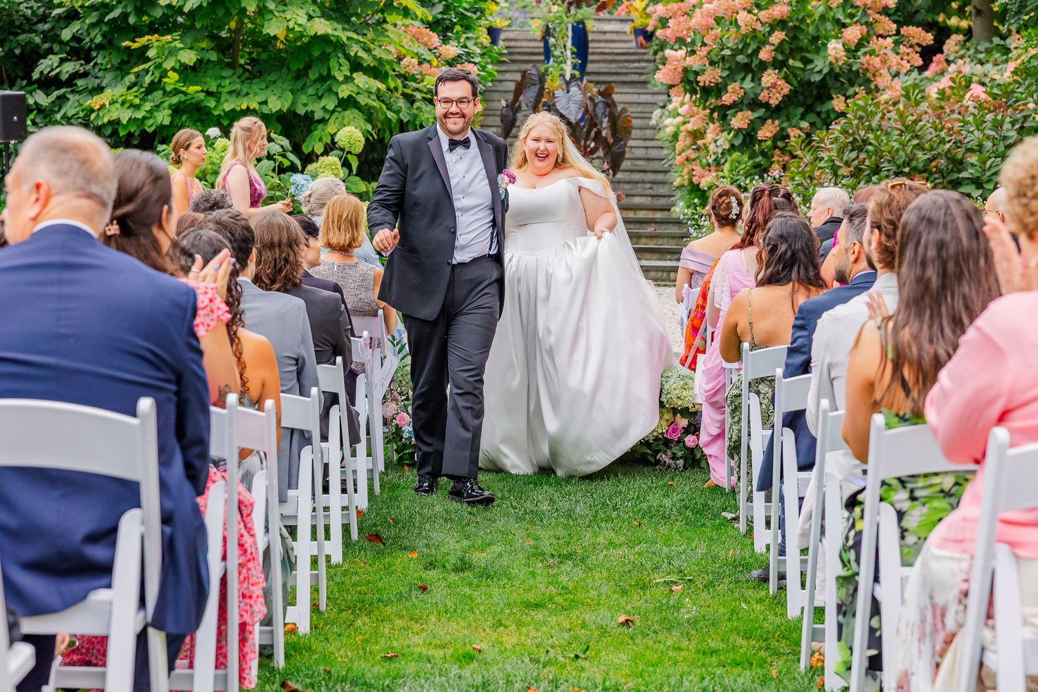Wedding guests seated in white chairs on a grassy lawn during an outdoor garden ceremony.