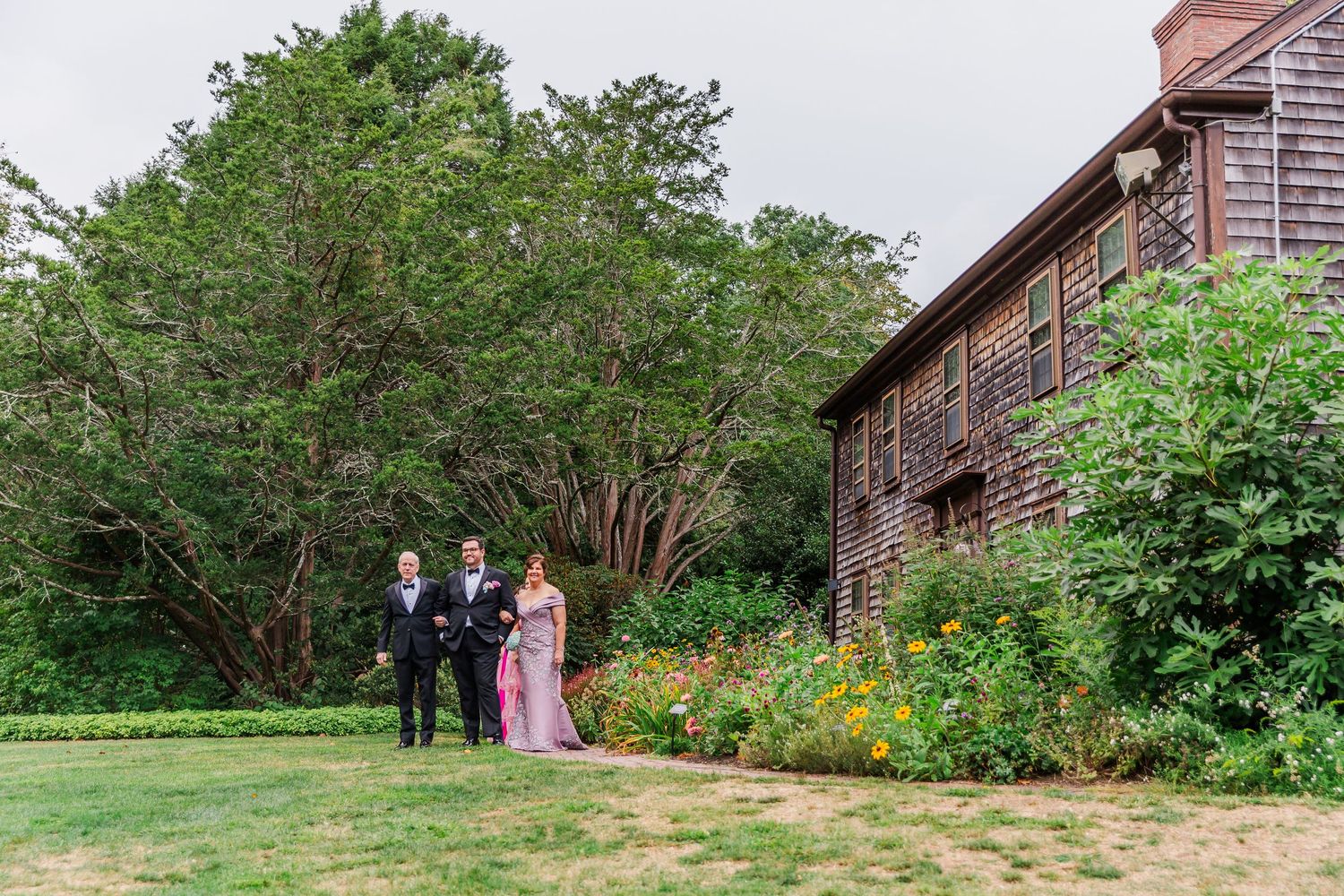 A group gathers in a lush garden setting with brick buildings and blooming flowers in the background.
