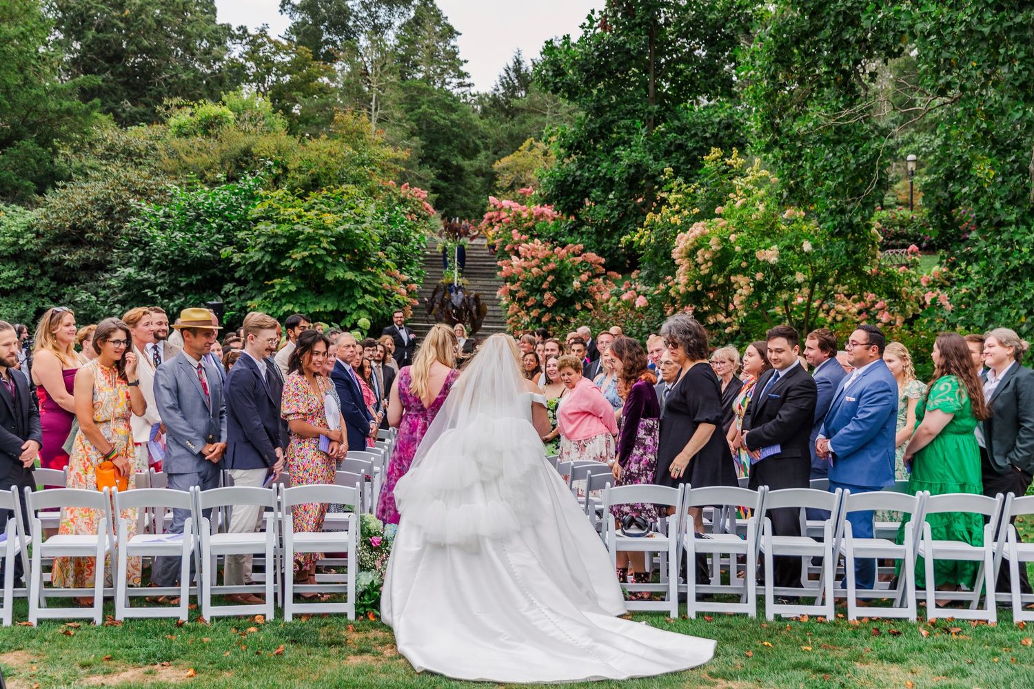 A group in formal attire poses along a lush garden pathway with flowering plants and trees.