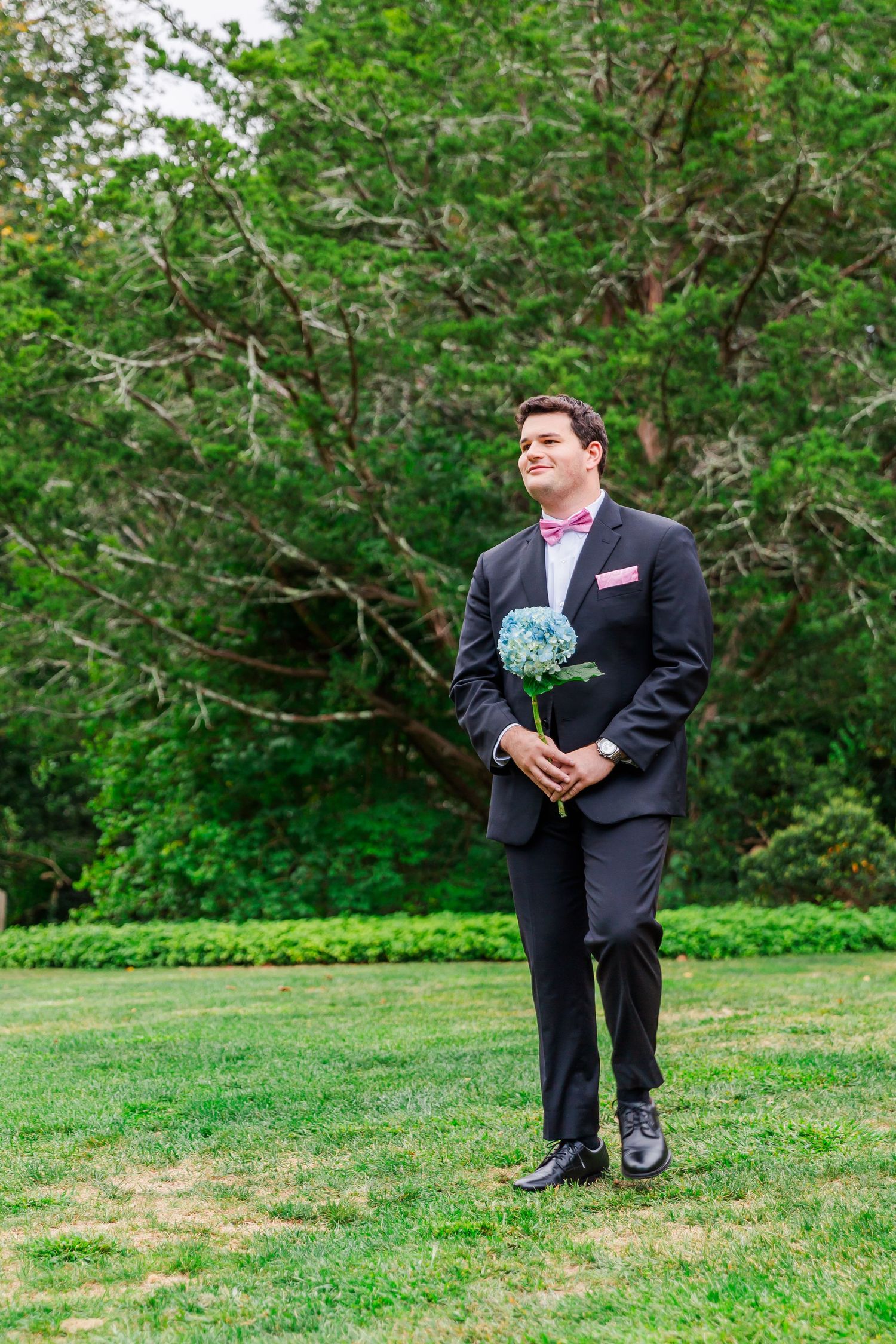 Someone in a dark suit stands on a green lawn holding a bouquet against a backdrop of lush trees and bushes.