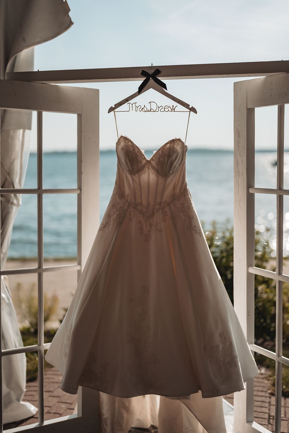 White wedding dress hanging in a window frame with ocean view in the background.
