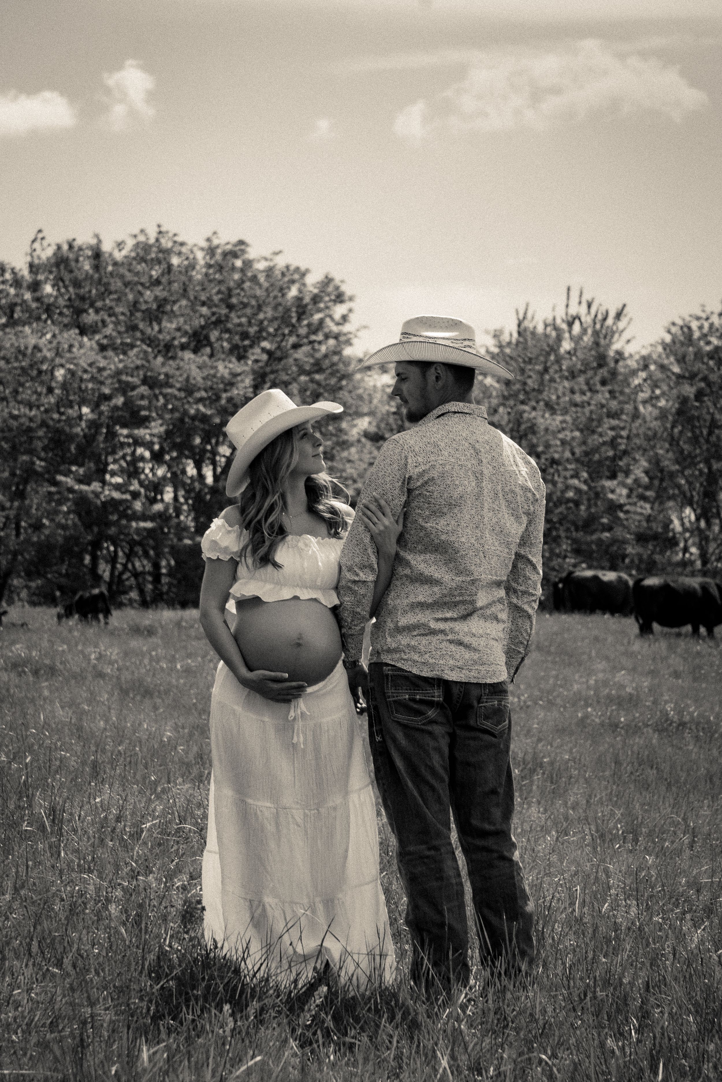 A black and white photo of a couple emphasizing the lady’s pregnant belly, looking at each other with trees and cows behind them.
