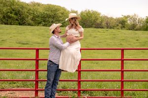 of a couple looking at each other. She’s sitting on a red gate and he’s leaning against it, looking up at her lovingly.