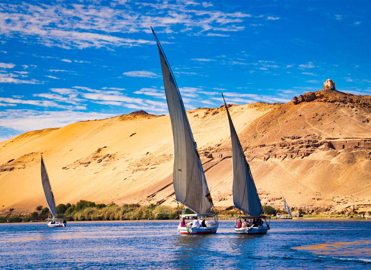 Traditional wooden sailboats glide along the Nile River with sandy desert hills and blue sky in the background.