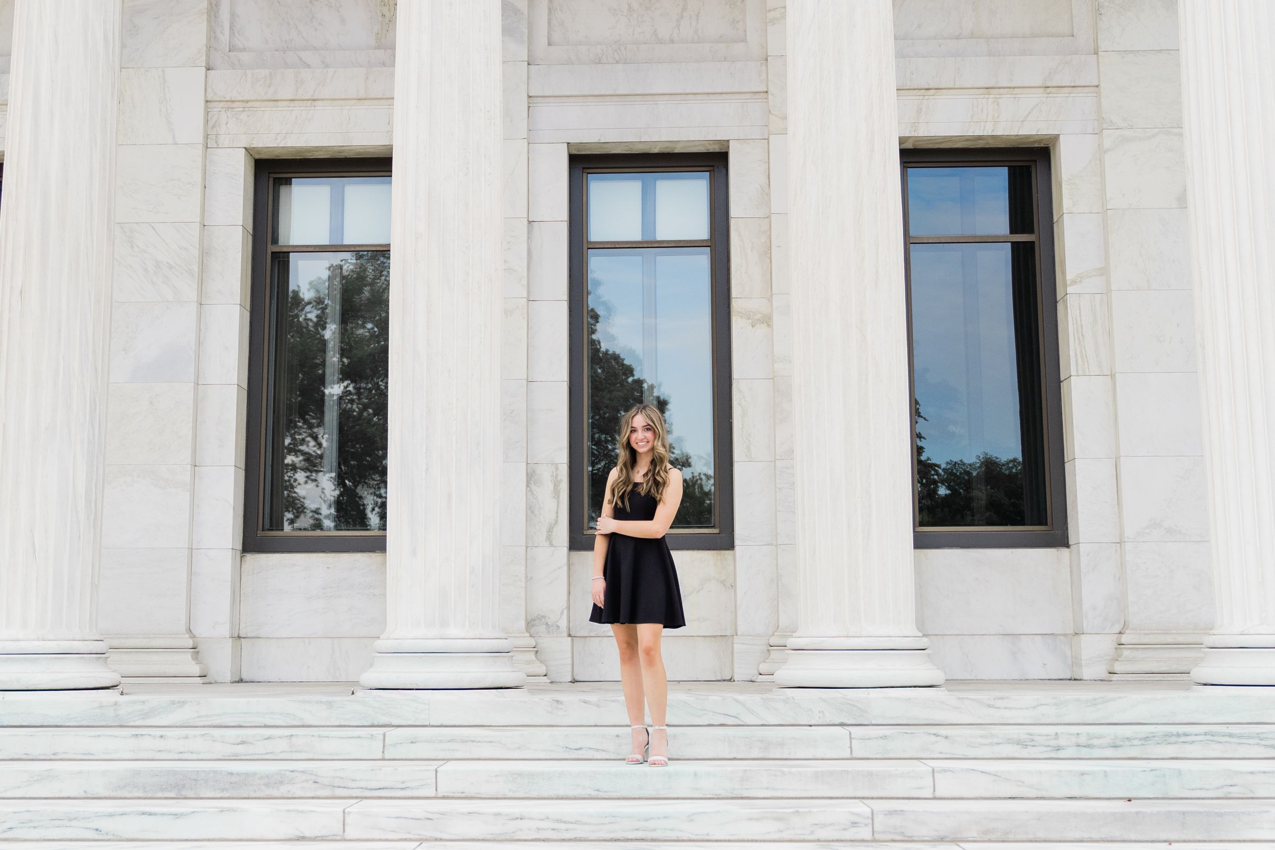 A woman in a black dress poses against a white marble building with classical columns and windows.