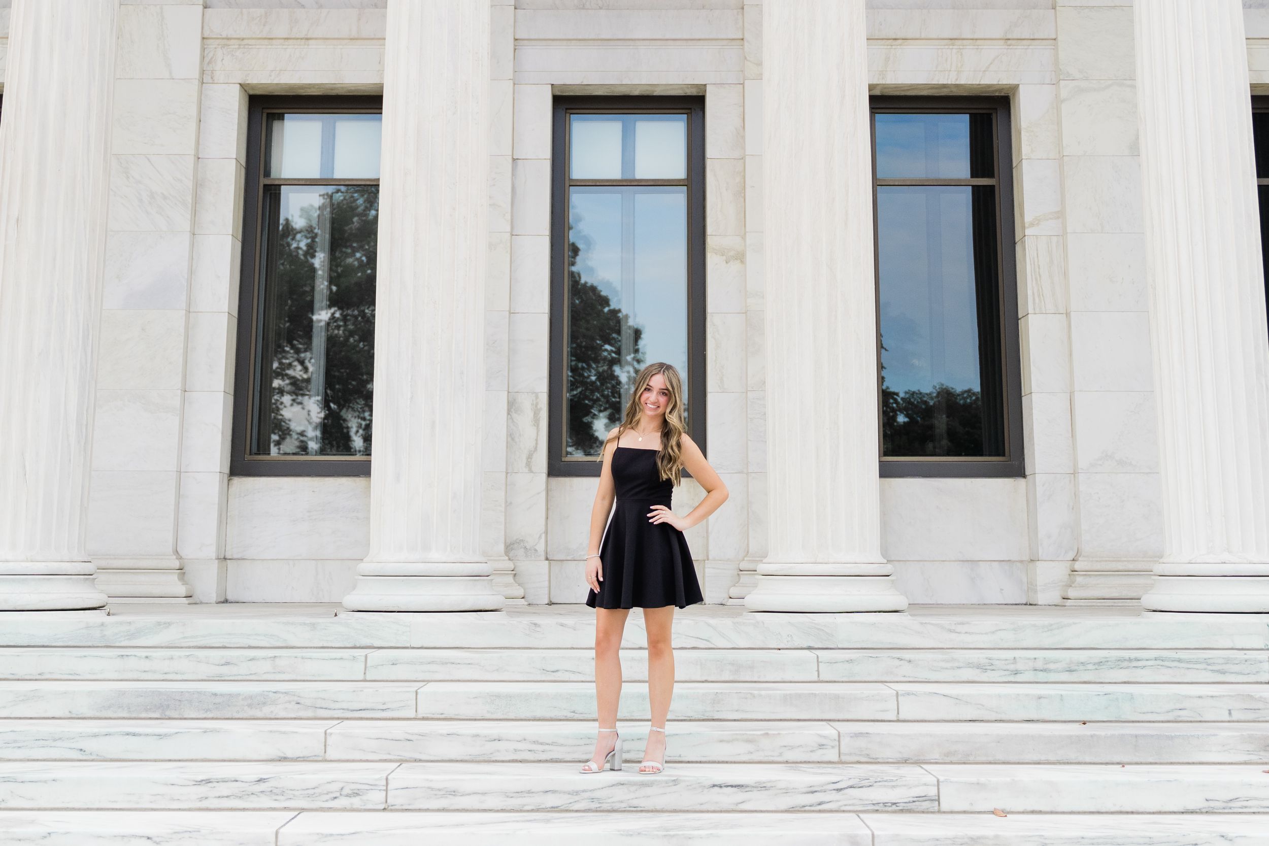 A group of graduates in black caps and gowns pose for photos on white stone steps of a grand building.