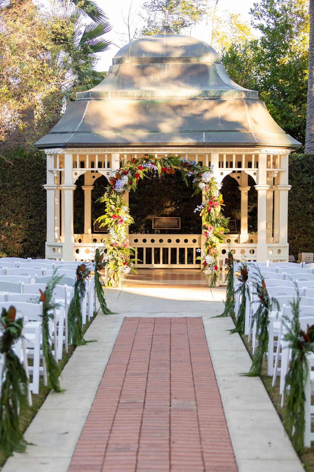 White wedding gazebo decorated with floral arrangements and greenery along a brick pathway at an outdoor venue.