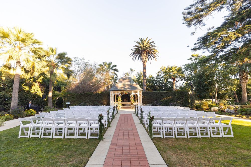 Panoramic view of a garden wedding ceremony setup with white chairs and a gazebo along a brick pathway at sunset.