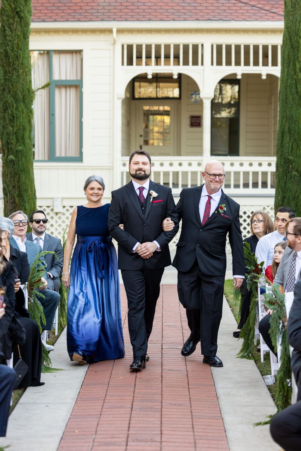 Wedding party members in navy dresses and black suits walk down a brick pathway during an outdoor ceremony.